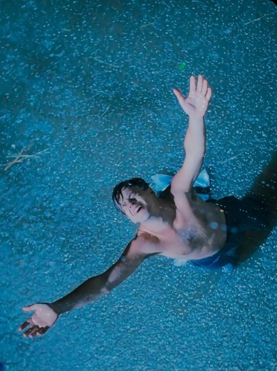 Still color photo from Shawshank Redemption of Andy Dufresne, after breaking out of the prison. He is shirtless. The water pools around him as he kneels drenched in rain with his arms outstretched to the sky. 