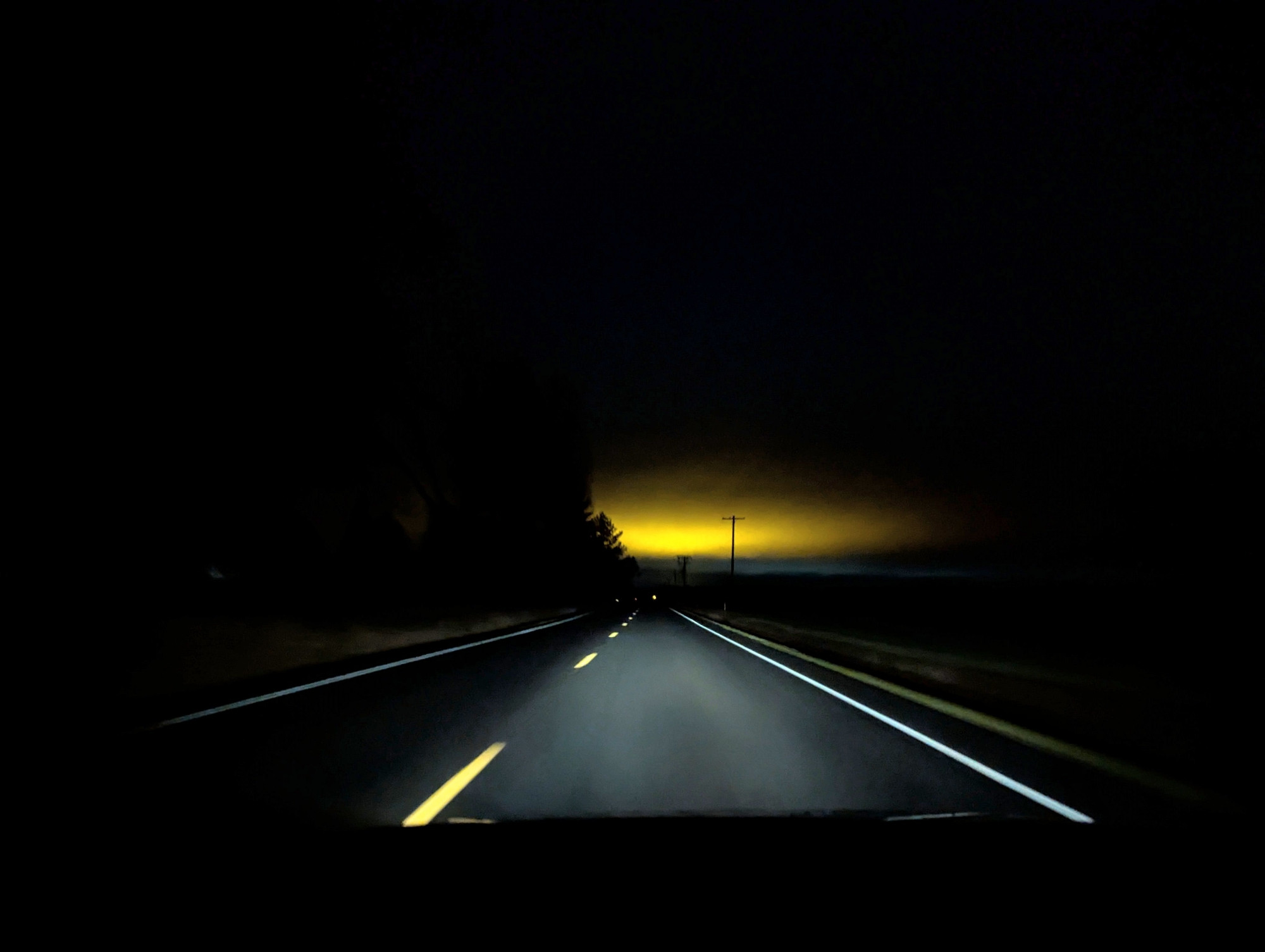 A rural, county road at night, the road and the American lane markings are lit by headlights, while in the distance low, overcast clouds are lit up by amber light pollution. There are trees occluding the light pollution to the left of the road, and some telephone poles silhouetting against it on the right side of the road.