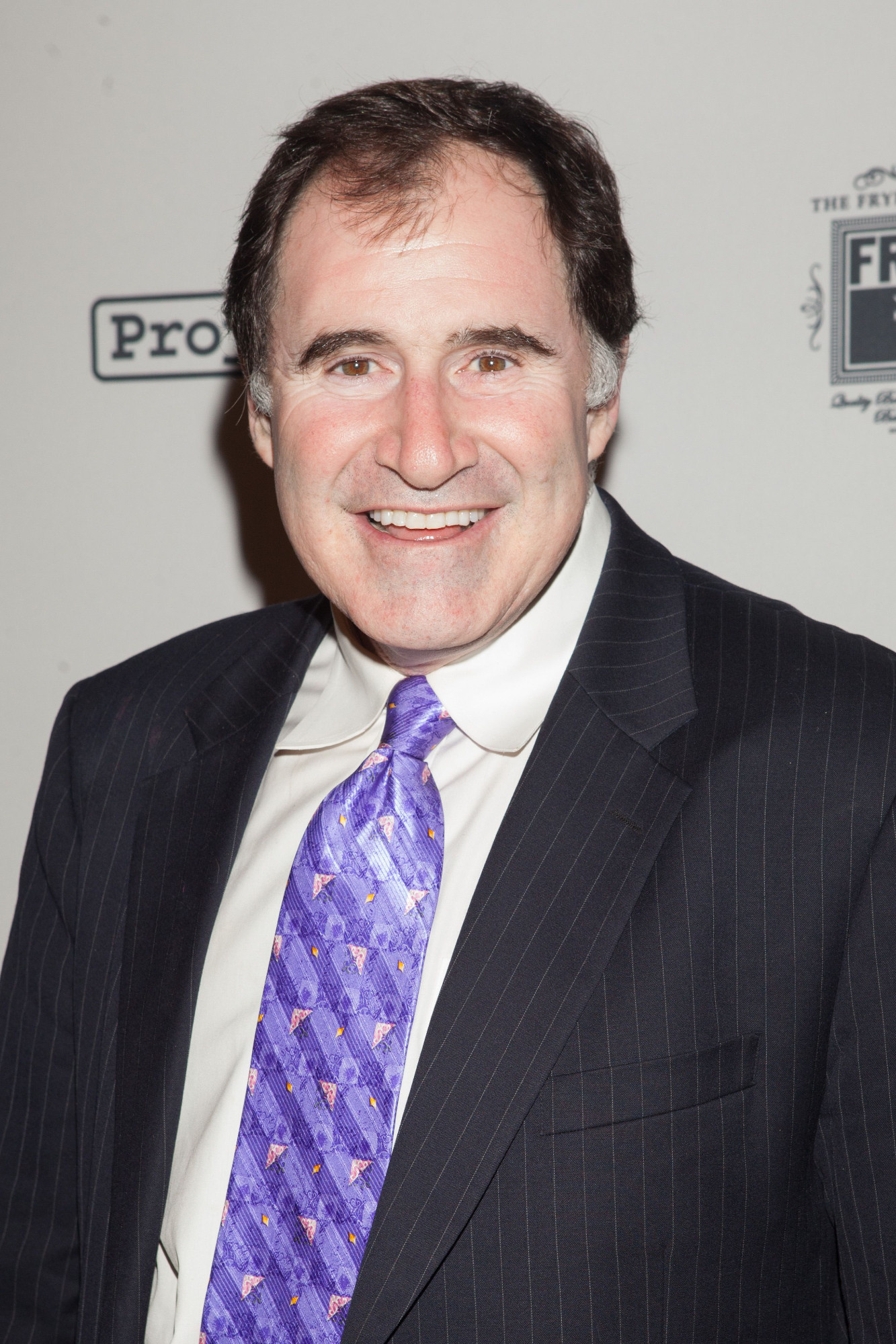 Photo of Richard Kind. A man in a black suit jacket, a white shirt, and a purple tie.