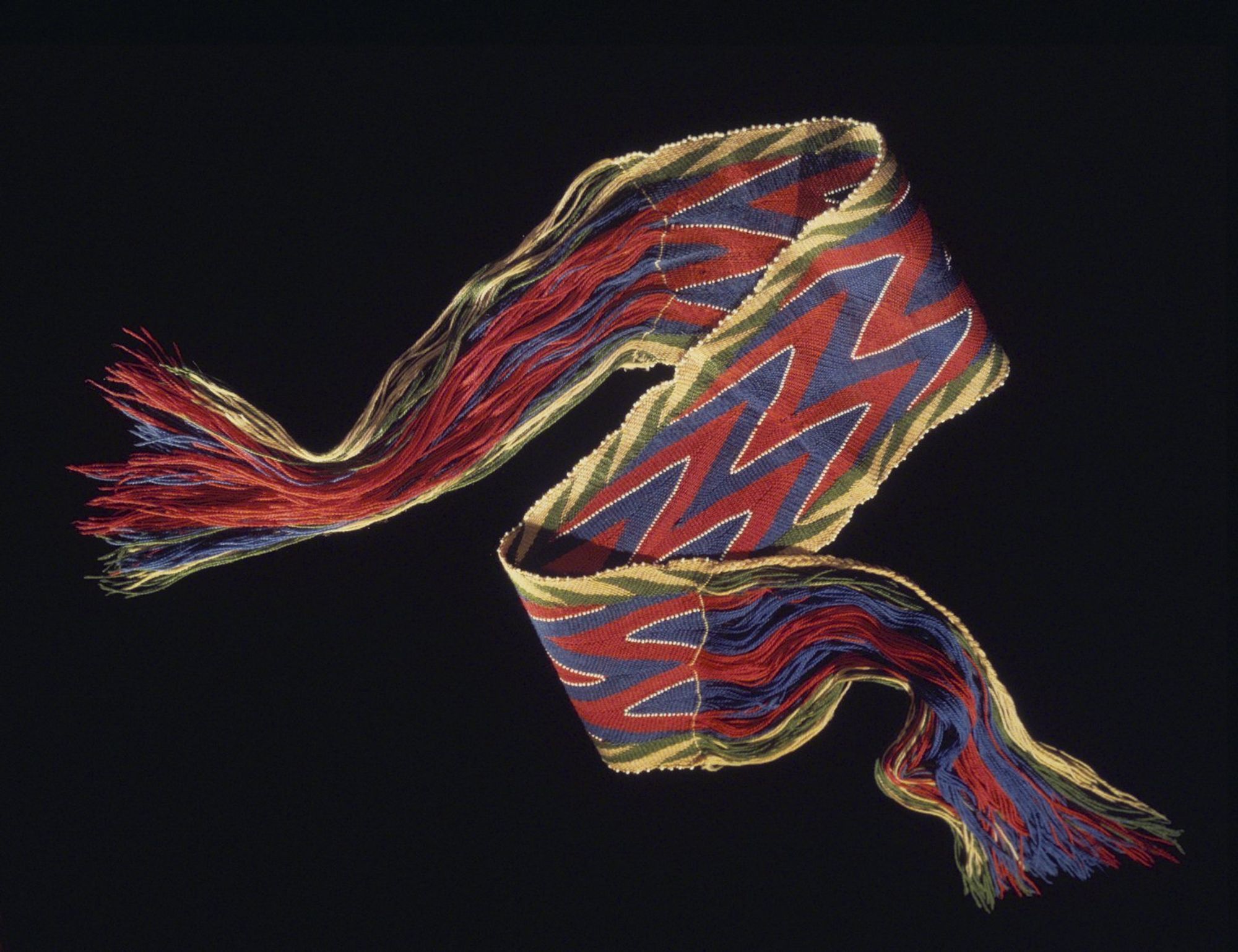 This is a finger  woven sash in bright colors of green, red, blue, white  and gold making a zig zag esign.  The ends are long fringes; the edges are trimmed with beads.