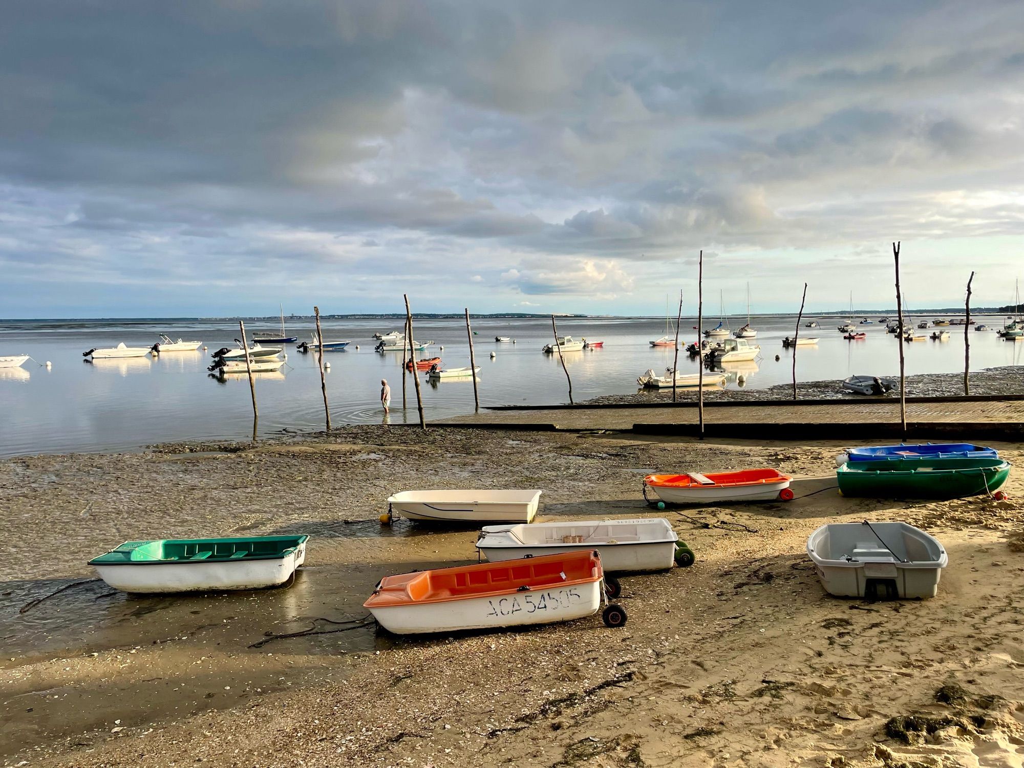 Marrée basse sur une plage du Cap Ferret, des bateaux en premier plan sont posés sur le sable. Tandis que les bateaux en arrière plan sont eux encore sur l’eau. Le ciel est un peu chargé en nuage.