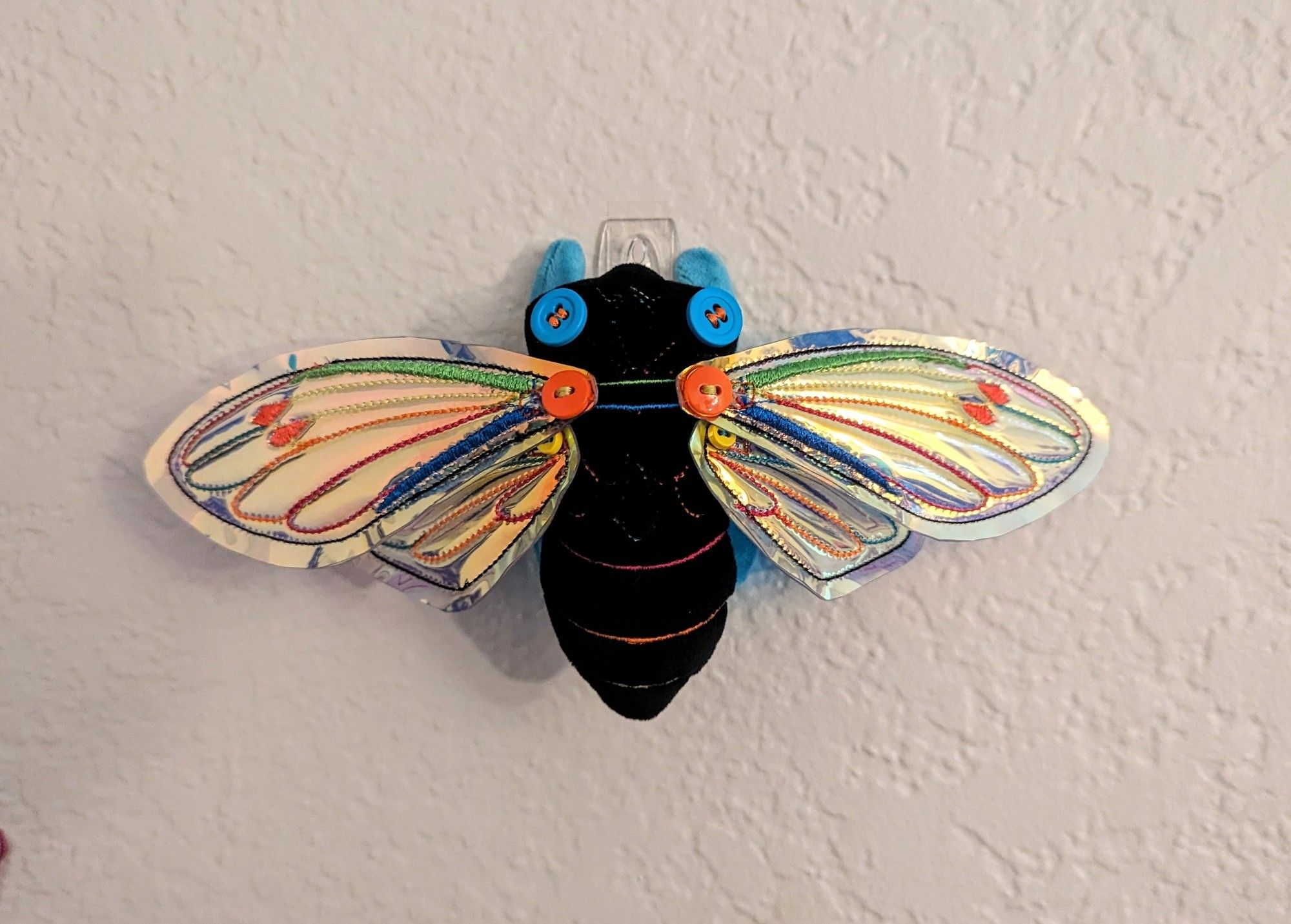 Image 3: The cicada art doll is hanging on a clear wall hook against a beige colored wall. The vinyl wings are shiny and reflective. The neon embroidery stands out against the black mochi minky and the iridescent vinyl.