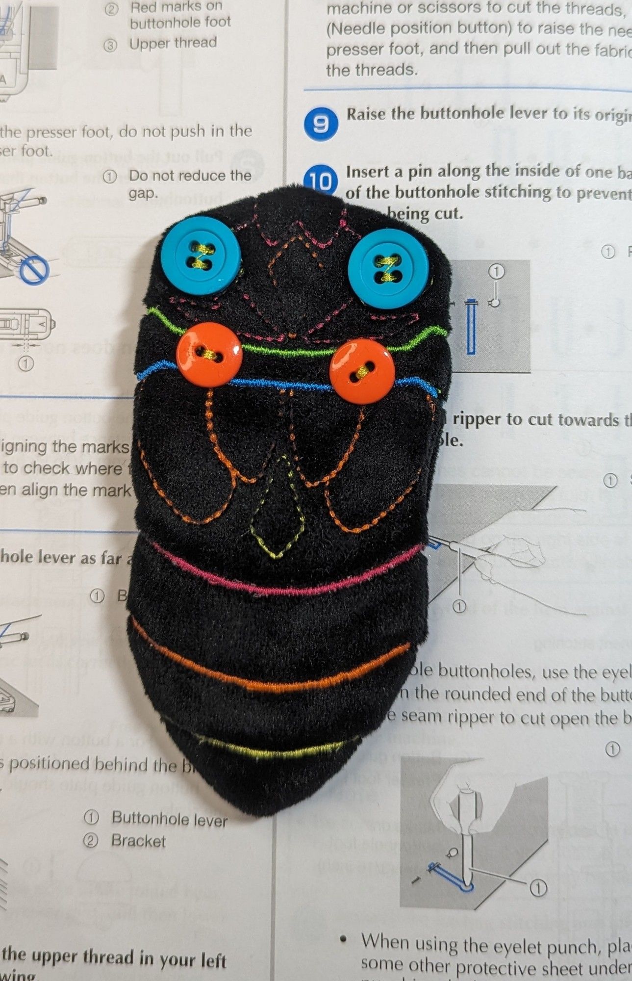 The body of a plush cicada.  The fabric is a velvet black and the pattern is embroidered with neon thread.  The cicada's eyes are two bright teal buttons.  There are two smaller orange buttons sewn on where the wings will be attached.