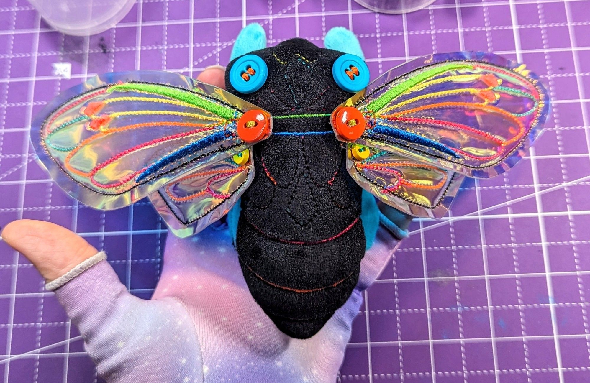 Image 1 - A plush cicada art doll with a black mochi minky body and neon embroidered details being held by a white person's hand wearing a pastel galaxy fingerless compression glove.  The wings are a shiny and opalescent vinyl with matching neon embroidery to the body. The wings are secured with orange and yellow buttons. The eyes of the cicada are bright teal buttons secured with neon yellow thread. The legs are black and teal mochi minky with armature wire to give them form.