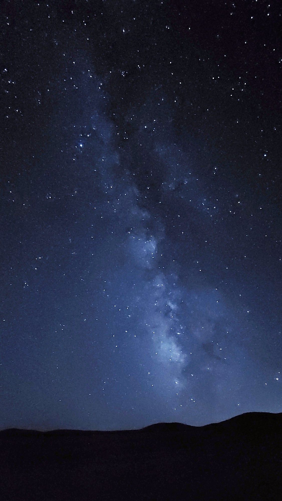 Night sky showcasing the Milky Way galaxy with numerous stars visible, seen above a silhouette of gentle hills.