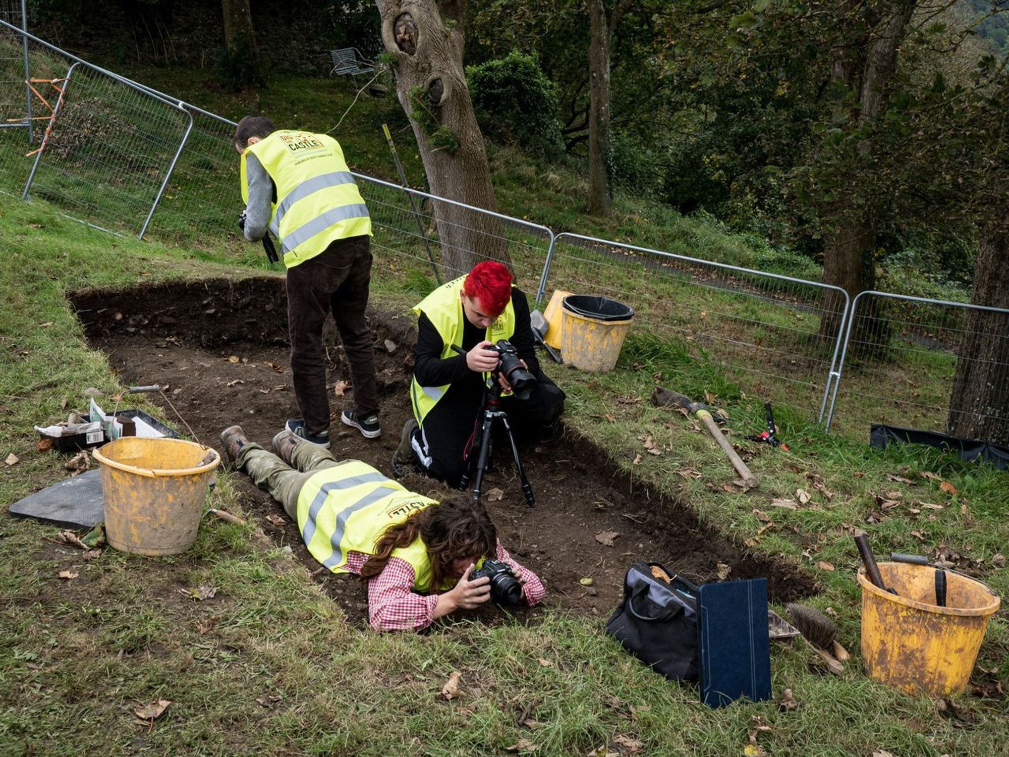 An archaeological trench approximately 4 metres long and 2 metres wide. There are three people in the trench, taking photographs with digital cameras. One person is lying down, one is kneeling and the other is standing up.