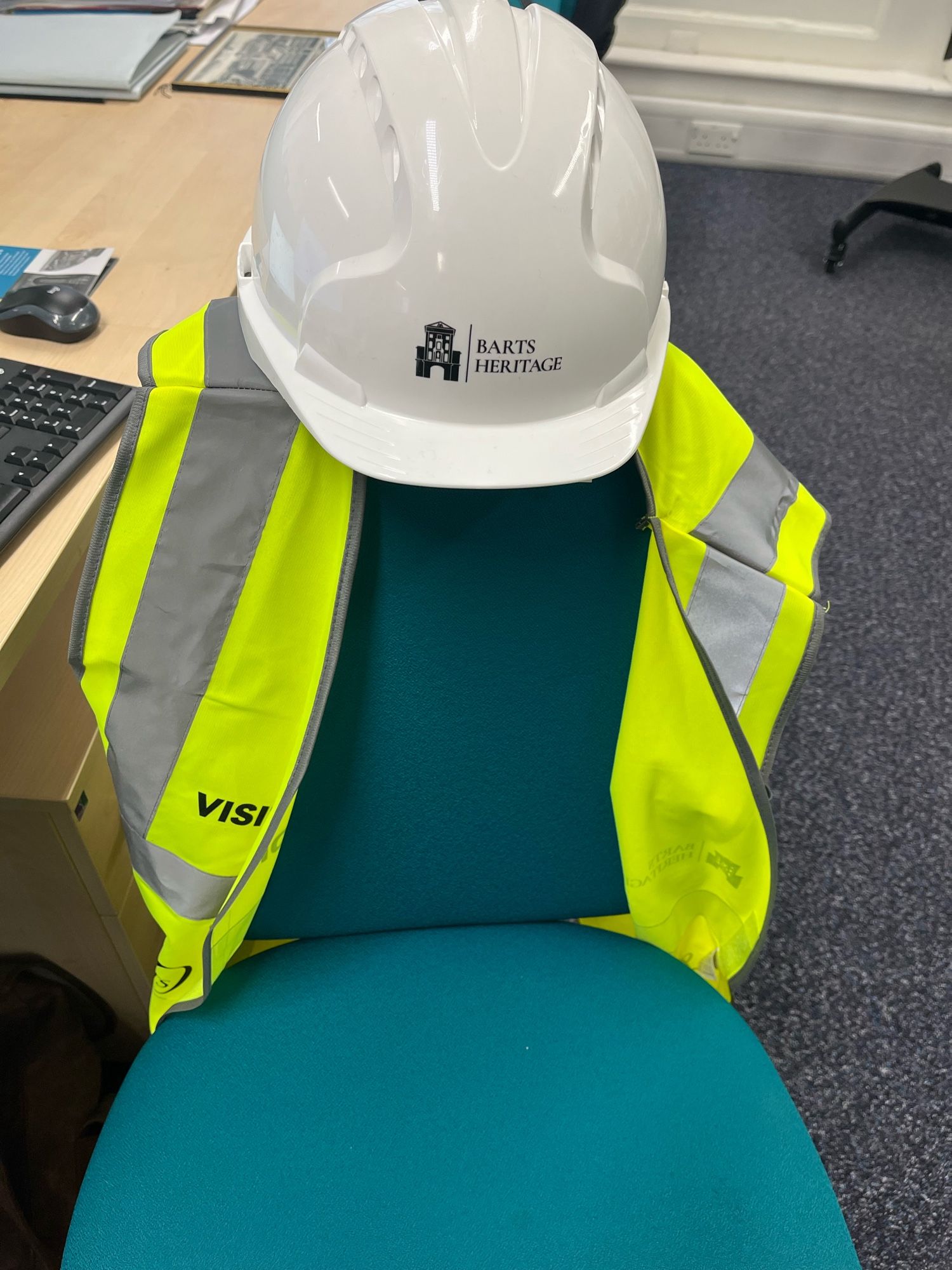 A white hard hat perched on an office chair with the logo Barts heritage on it. A high vis jacket is hung on the chair