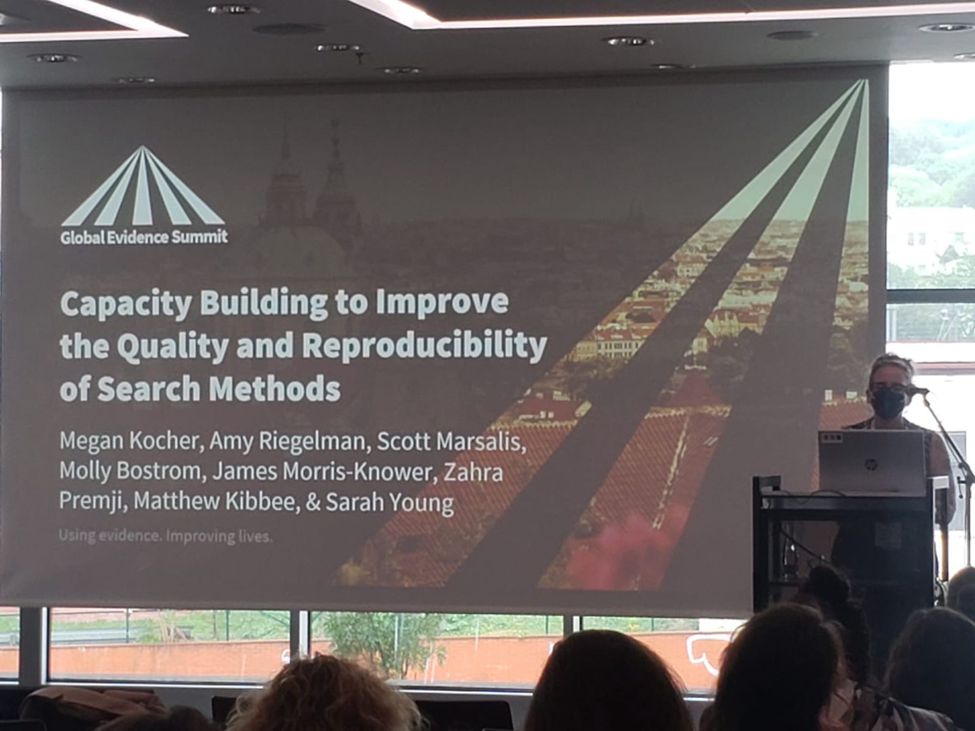 Masked speaker (me) in front of a screen onto which is projected a slide titled Capacity Building to Improve the Quality and Reproducibility of Search Methods