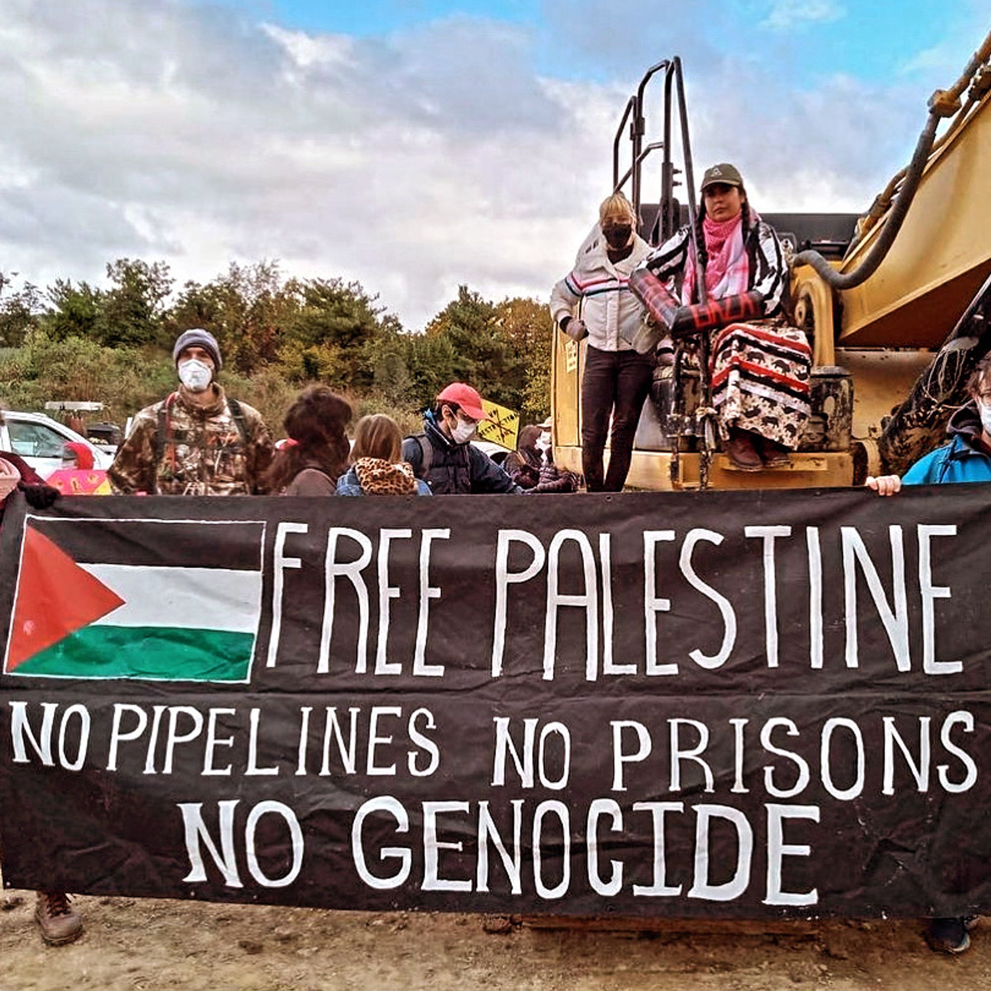 Banner reads Free Palestine. No Pipelines. No Prisons.No Genocide. A person with a ribbon dress is locked to an excavator