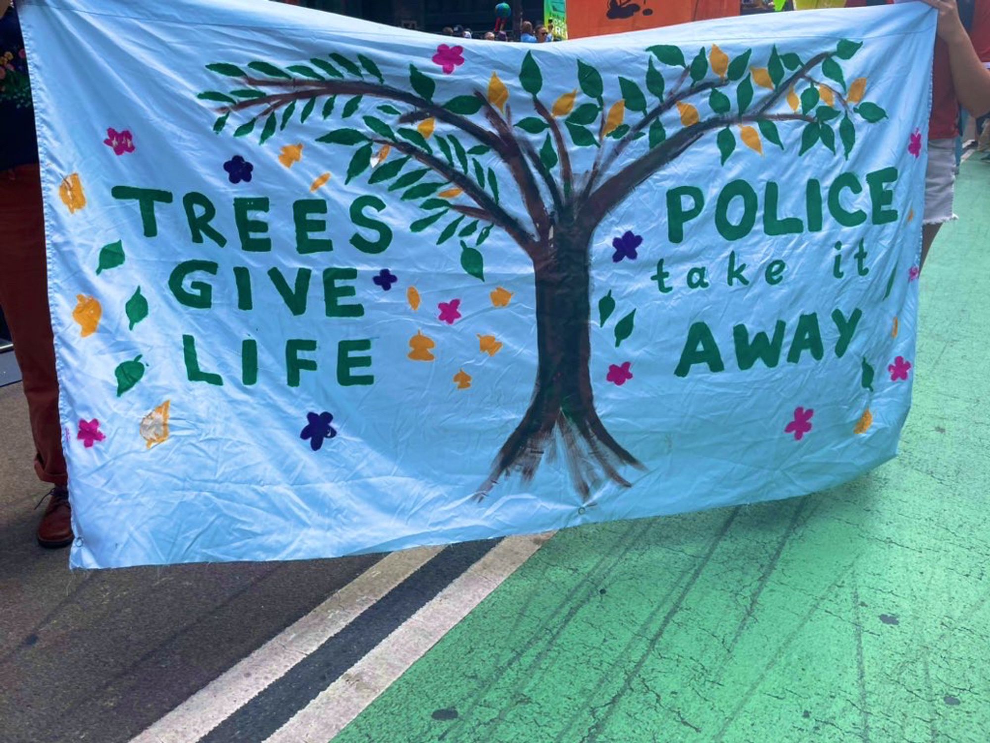 A light blue banner with a beautiful tree painted in the middle reads, “Trees give life, police take it away” leaves surround the whole banner.