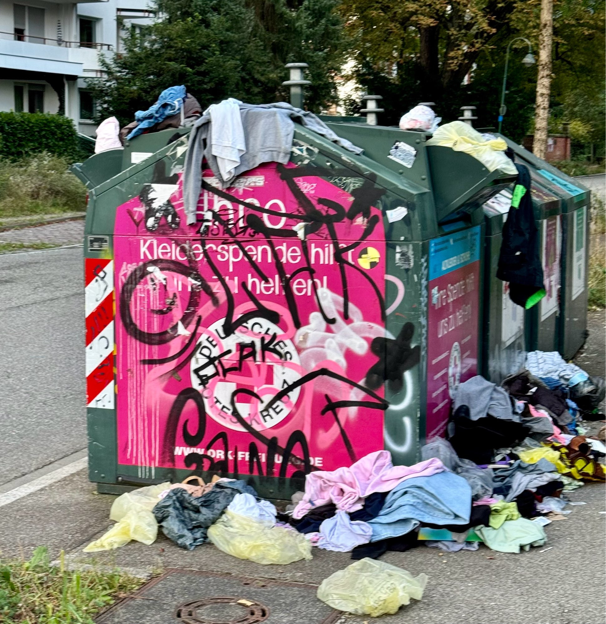 Kleidercontainer, aus dem viele Kleider herausgerissen und daneben auf Haufen geworfen wurden.