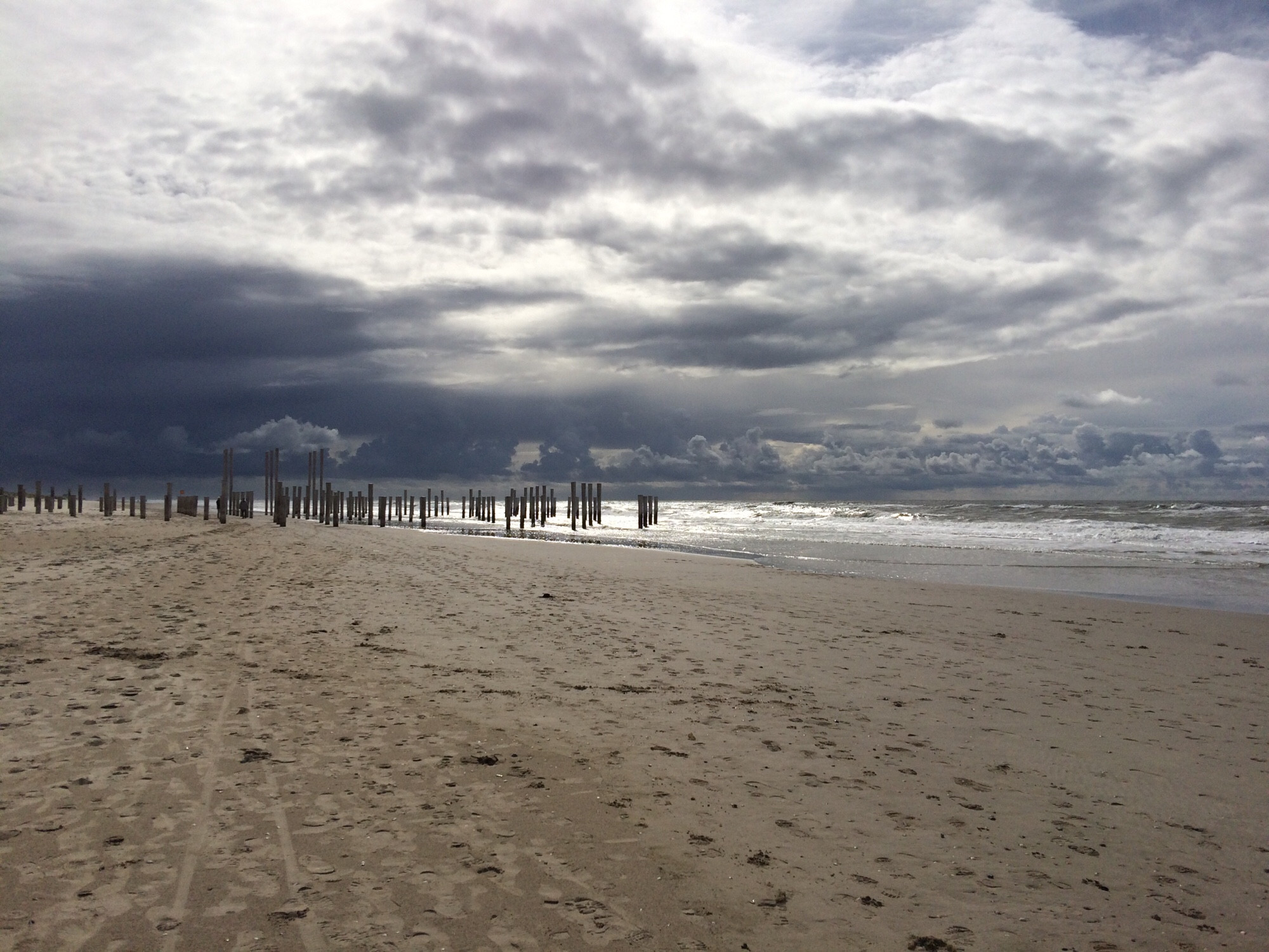 Herfstfoto van een verlaten strand. Petten aan Zee, september 2017. Op de voorgrond een grote zandvlakte met sporen van mensen en honden. Midden in beeld een grote verzameling strandpalen (minstens 50) die als een soort groep toeschouwers op het strand en in zee staan (deels). Er zijn een aantal (9) palen die erg hoog zijn; de hoge palen zijn ongeveer dubbel zo hoog als de gewone strandpalen en ze staan in 3 rijen van 3 palen. De zilvergrijze zee is rechts in beeld en is ook achter de palen die in zee staan te zien. De zee ziet er redelijk rustig uit, de golven zilverwit. De bovenste helft van het beeld is behoorlijk donker: er hangt een grote, bijna zwarte wolk boven het strand die de lucht wat onheilspellends geeft. Boven zee en helemaal bovenaan in beeld is de lucht echter veel lichter van kleur, er zijn wolken te zien die veel witter zijn. Ergens door de wolken heen valt er voldoende zonlicht op de zee om die zilverwit te doen oplichten. Het is echter beslist geen zonnige foto.