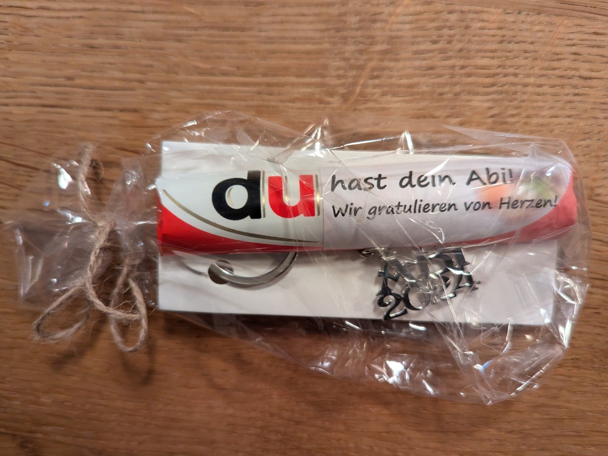 Ein Duplo mit einer Banderole, auf der "Du hast dein Abitur, wir gratulieren von Herzen " steht. Es liegt in einer Klarsichtfolie zusammen mit einem silbernen Schlüsselanhänger "Abitur 2024".