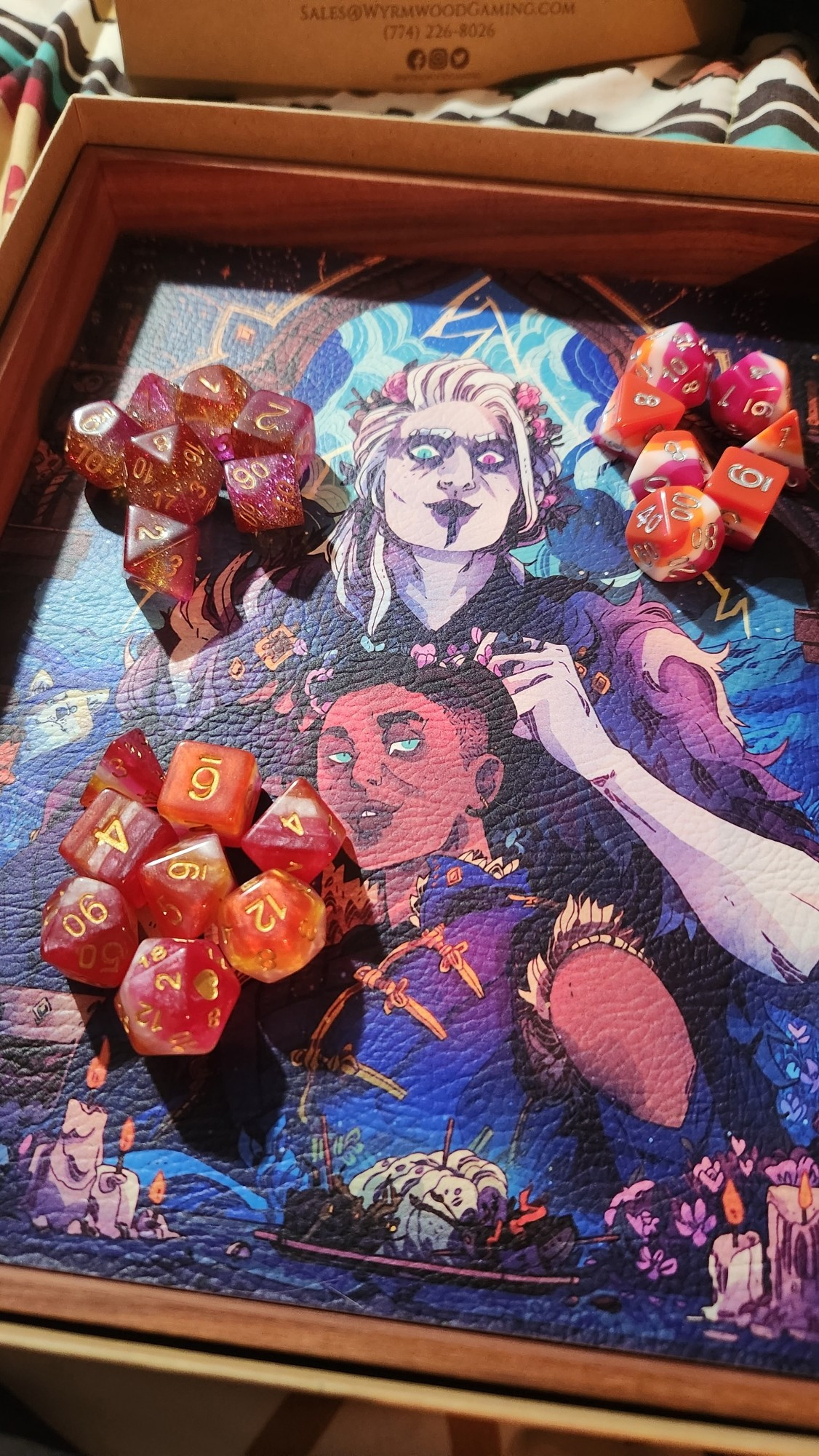 three sets of dice in the lesbian flag colors, one set is pastel, one has glitter, and one is opaque. all of the dice are on a wyrmwood dice tray that has beau and yasha from critical role as the art.