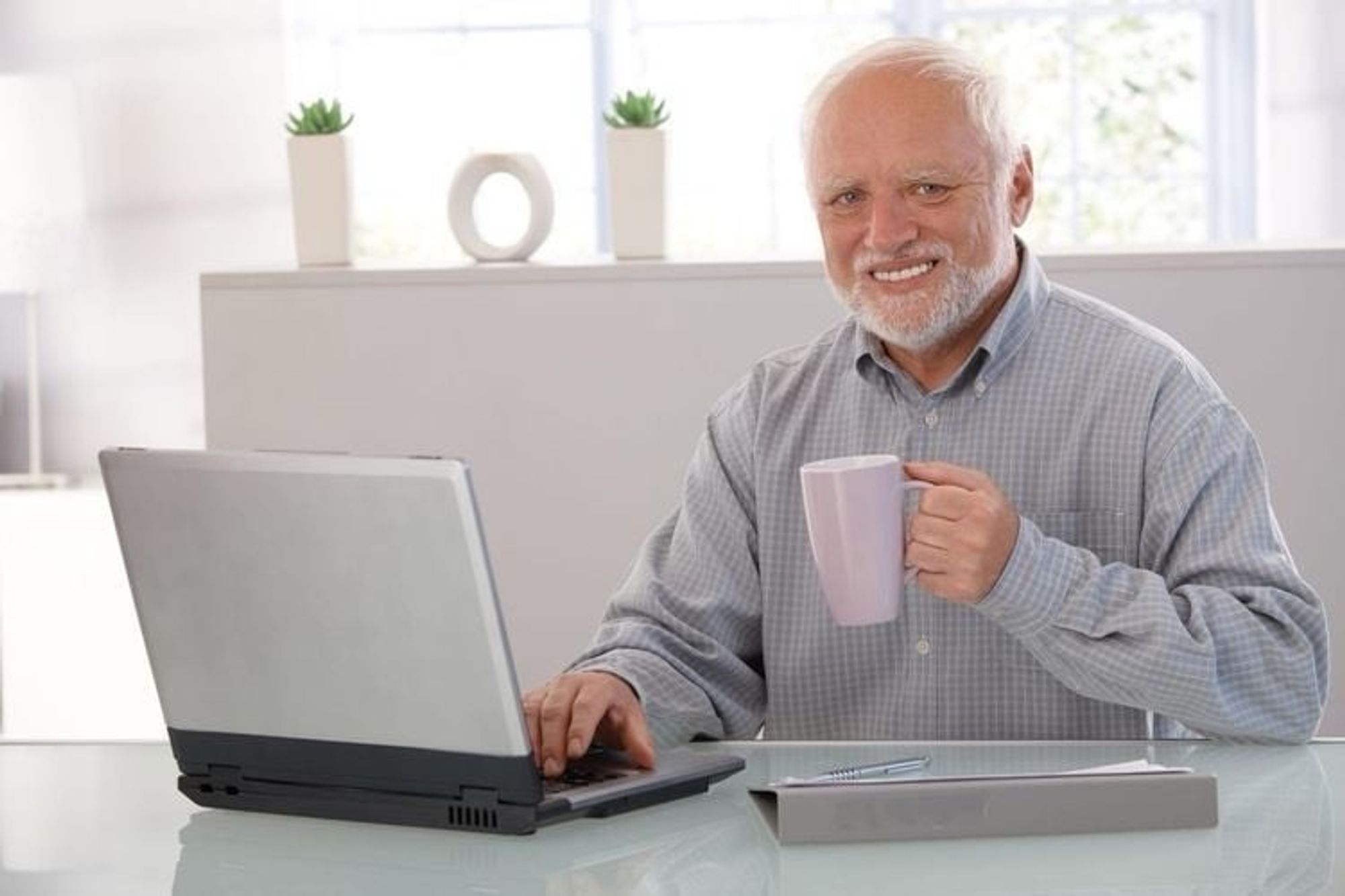 An older man with a gray beard smiles while sitting at a desk. He is holding a light purple mug and typing on a laptop. Small plants and a decorative item are visible in the background.