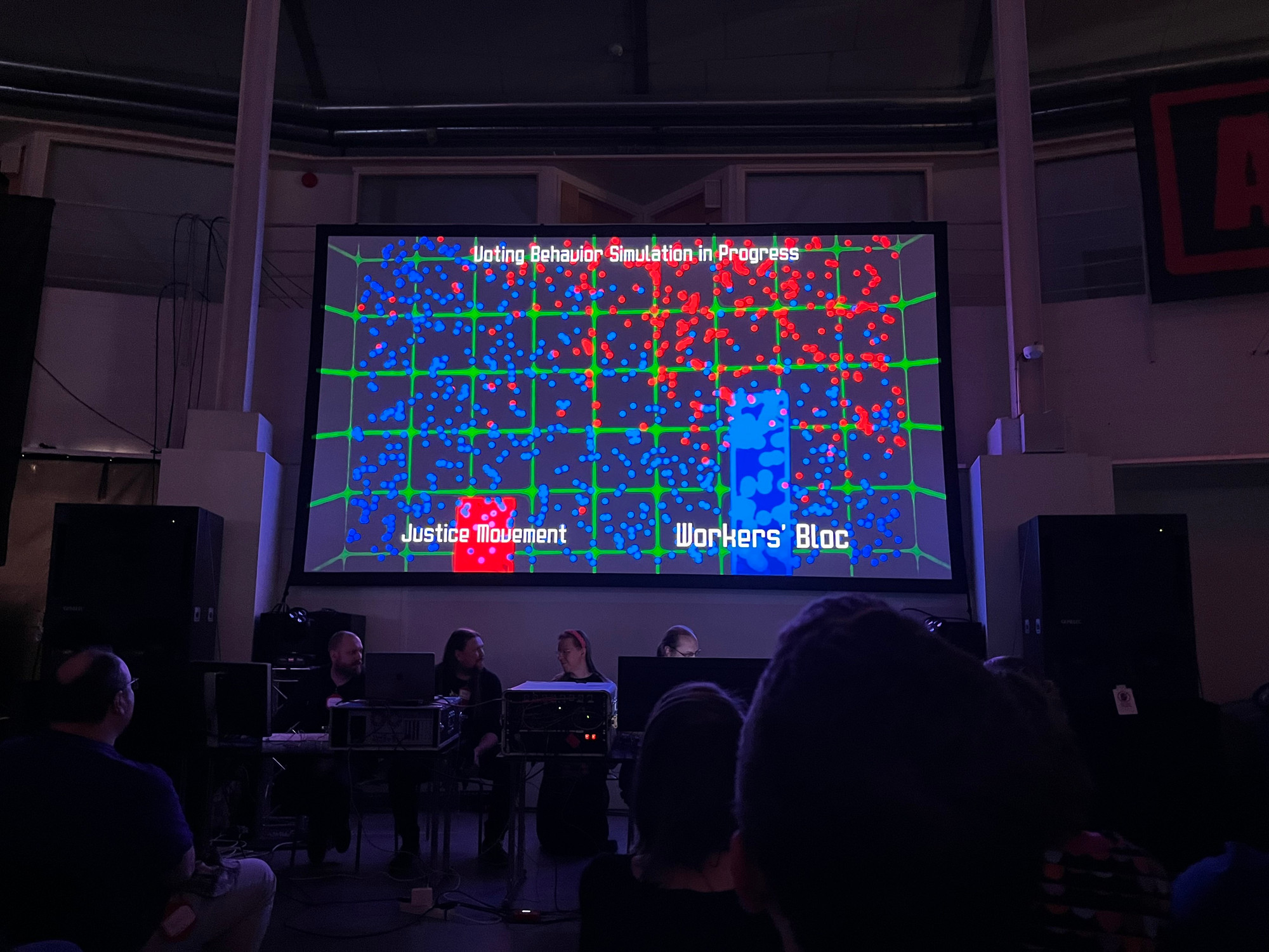 The image shows a presentation screen displaying a colorful simulation titled "Voting Behavior Simulation in Progress." It features a grid with blue and red dots, representing two groups: "Justice Movement" and "Workers' Bloc."