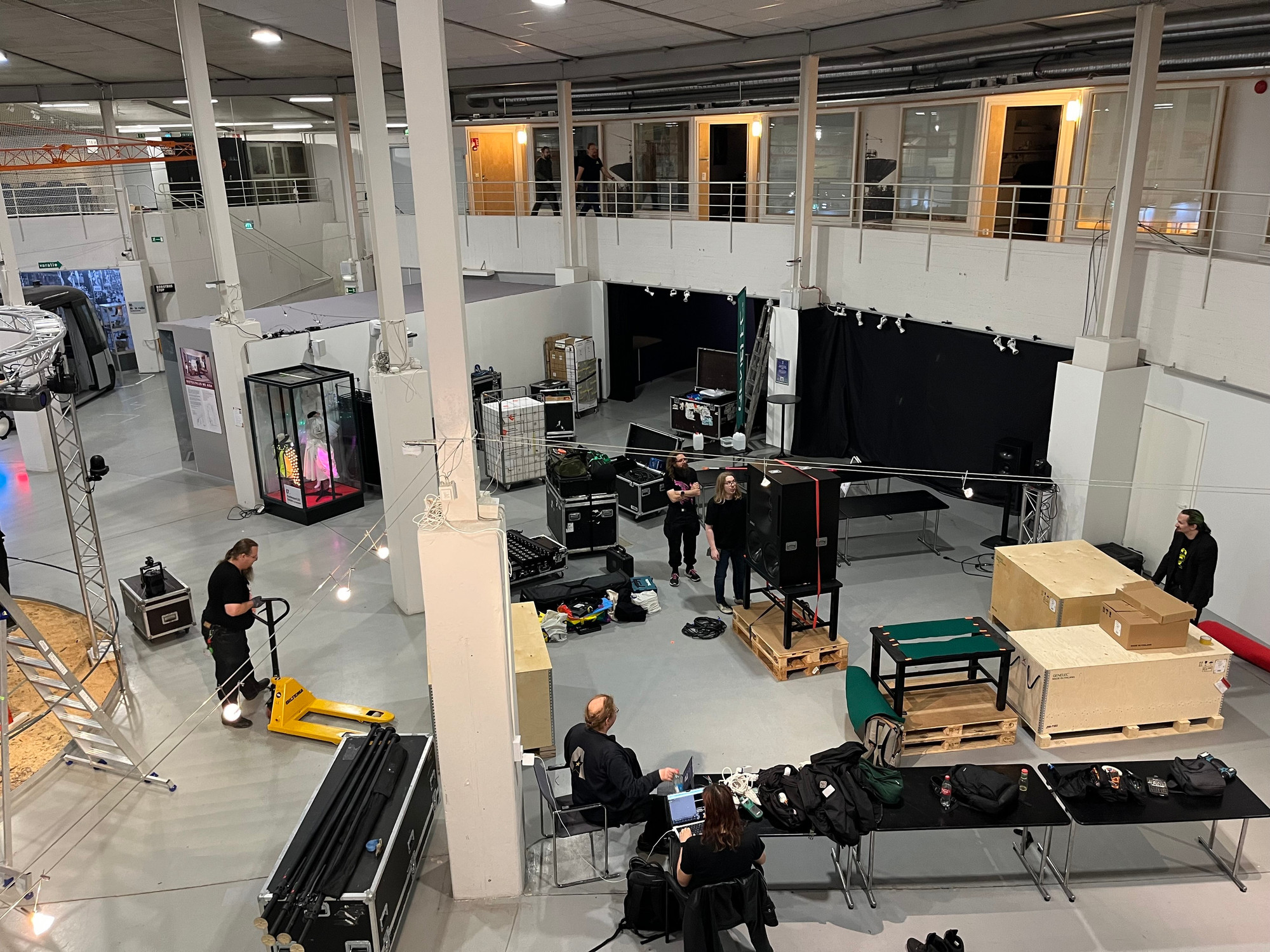 A spacious indoor venue with various setups, including equipment cases and pallets. The Alt Party crew can be seen preparing, with some working at tables and others moving items. A display case containing an illuminated object is visible. The environment features high ceilings and industrial look. 