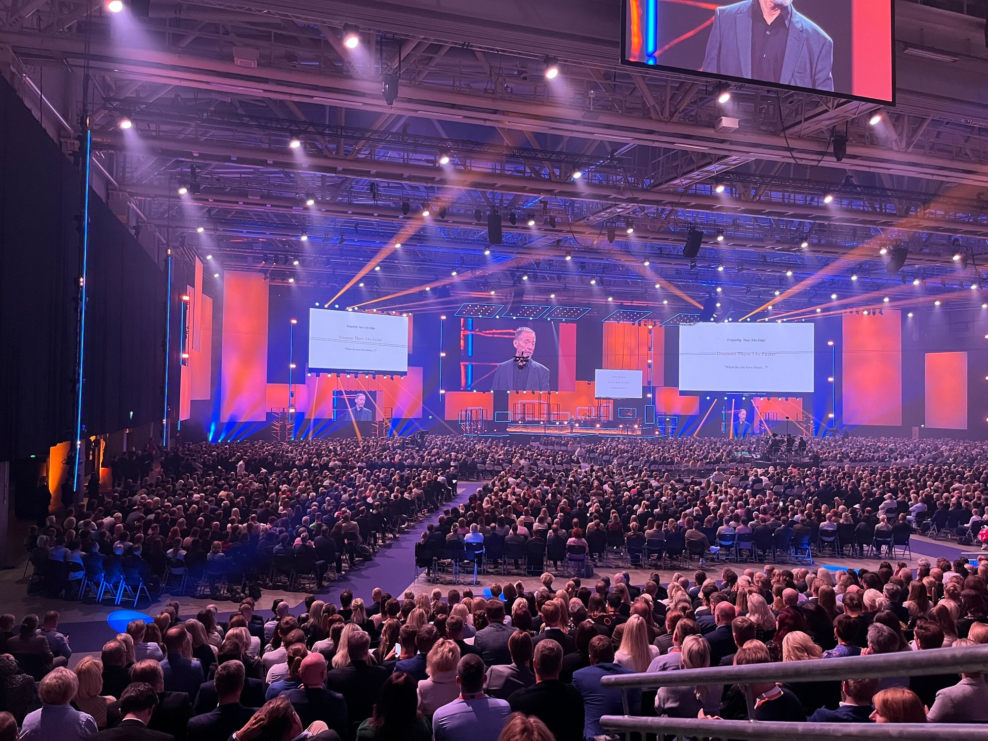 A large conference or seminar featuring a speaker on stage, with a packed audience seated in rows. The venue is illuminated with colorful lights, and projection screens are displaying text above the speaker.