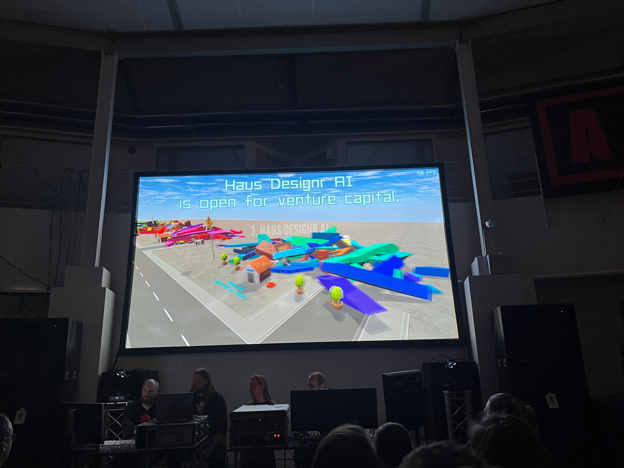 A digital presentation display shows a colorful 3D model of buildings and objects, along with the text "Haus Design AI is open for venture capital." In the foreground, a group of people is seated at a table with computers.