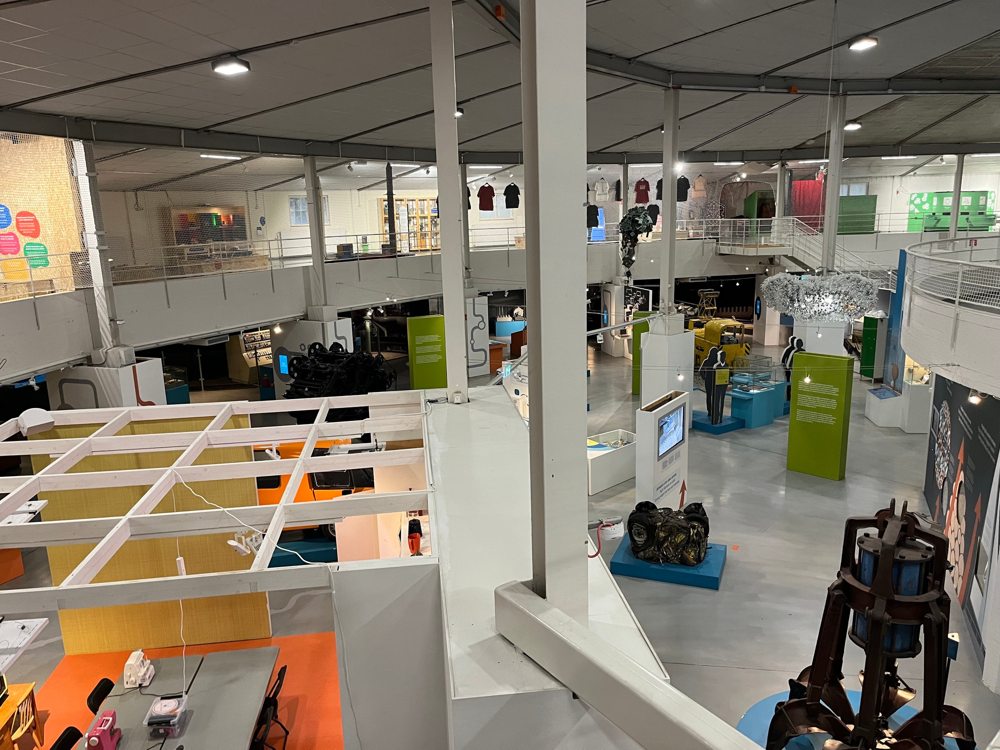 An aerial view of a modern exhibition space featuring various interactive displays and exhibits. The area includes tables, information panels, and displays with colorful graphics. Suspended artworks are visible.