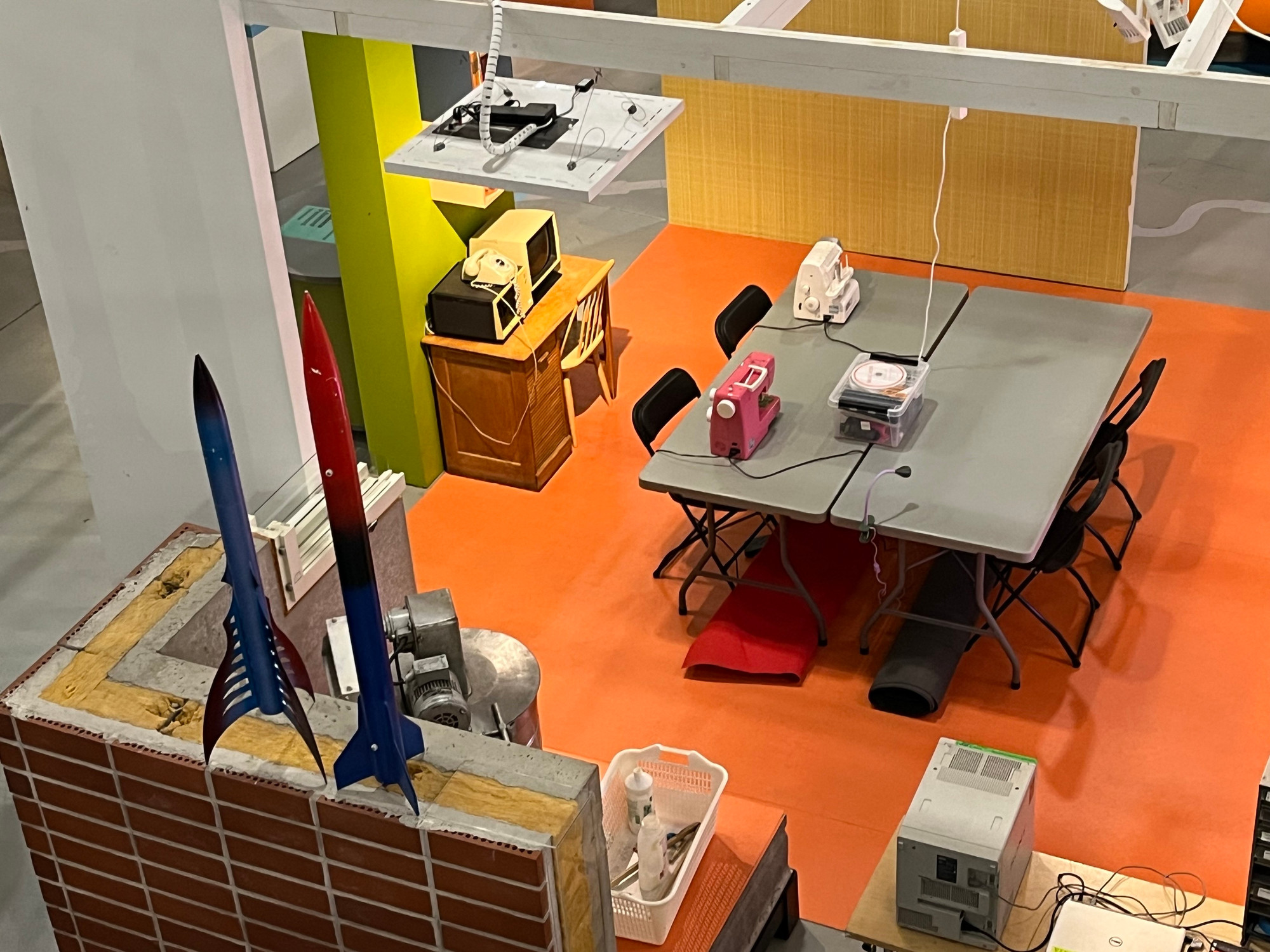 An overhead view of a creative workspace featuring a gray table surrounded by black chairs, sewing machines, and various electronics. Two colorful model rockets are displayed on a ledge, alongside a vintage desk and an old monitor. 