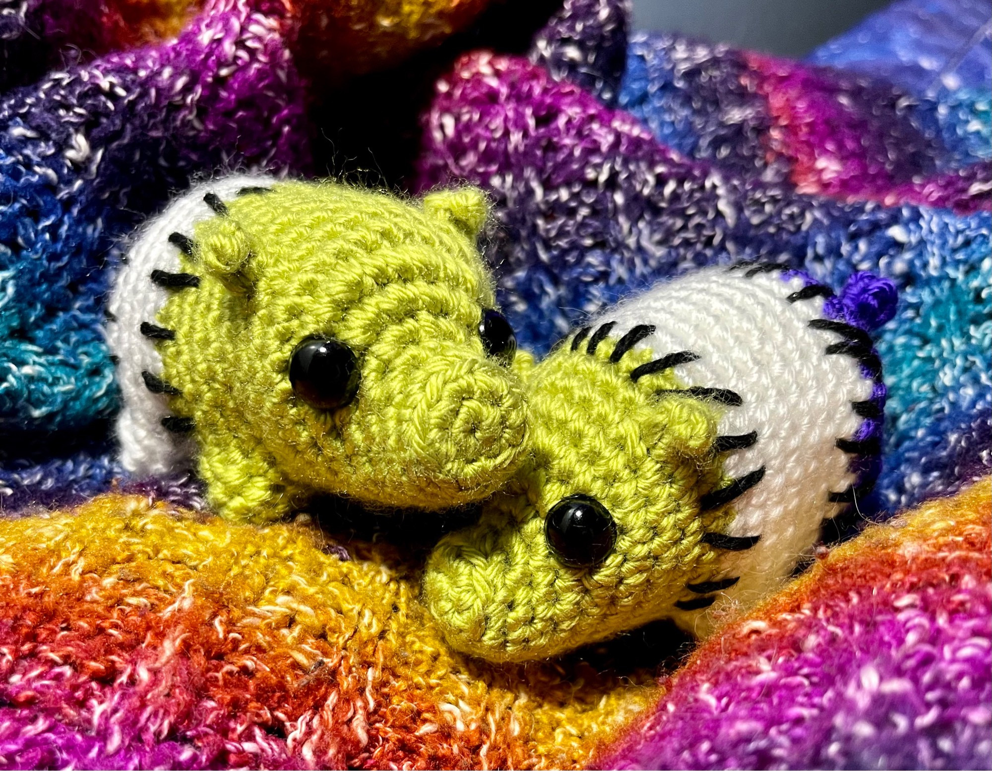 Two crochet pigs in green, white and purple. Each piece of pig is stitched together with big black stitches.