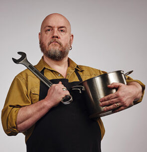 Snack hacker George Egg, a sturdy, bald man with a greying beard. He has a pan in one hand and a spanner over one shoulder.