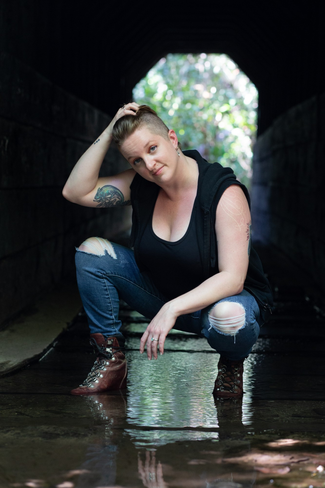 Author Sarah Gailey crouches in a stream. A tunnel is behind them with sunshine and trees visible in the distance. They're wearing boots, jeans and a black top.