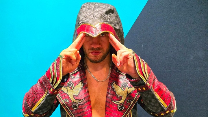 Wrestler Will Ospreay, dressed in a black red and gold assassin cowl, stares at the camera.