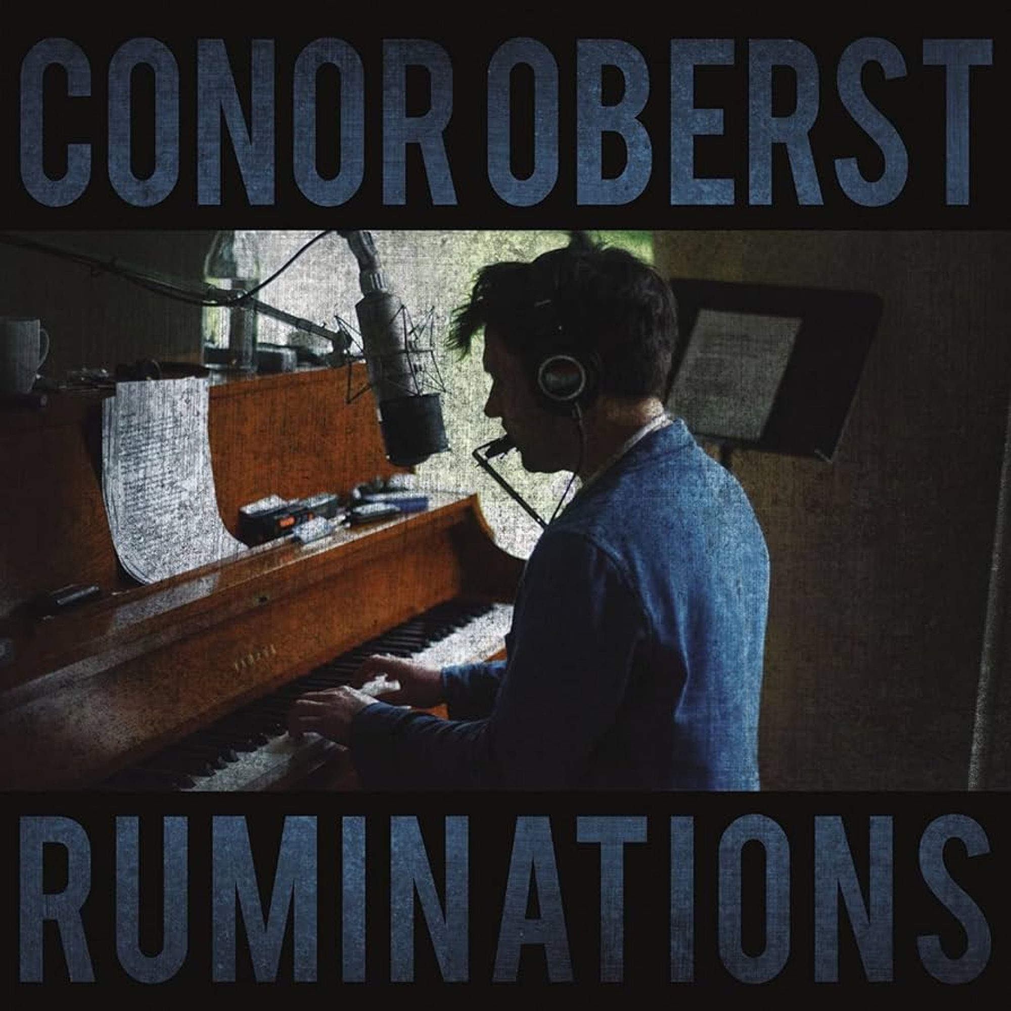 The image shows the album cover for Ruminations by Conor Oberst. The cover features a dark, intimate setting with Conor Oberst seated at a piano. He is captured in profile, wearing headphones and playing the piano, while also singing into a vintage-style microphone. There is a harmonica strapped around his neck, adding to the raw, acoustic feel of the scene. Papers, possibly lyrics or sheet music, rest on the piano. The lighting is dim and natural, emphasizing the solitary and reflective mood of the image. The artist's name "Conor Oberst" is written in large, bold letters at the top, and the album title "Ruminations" is displayed in similarly large text at the bottom, both in a deep blue hue that blends with the dark, textured background.