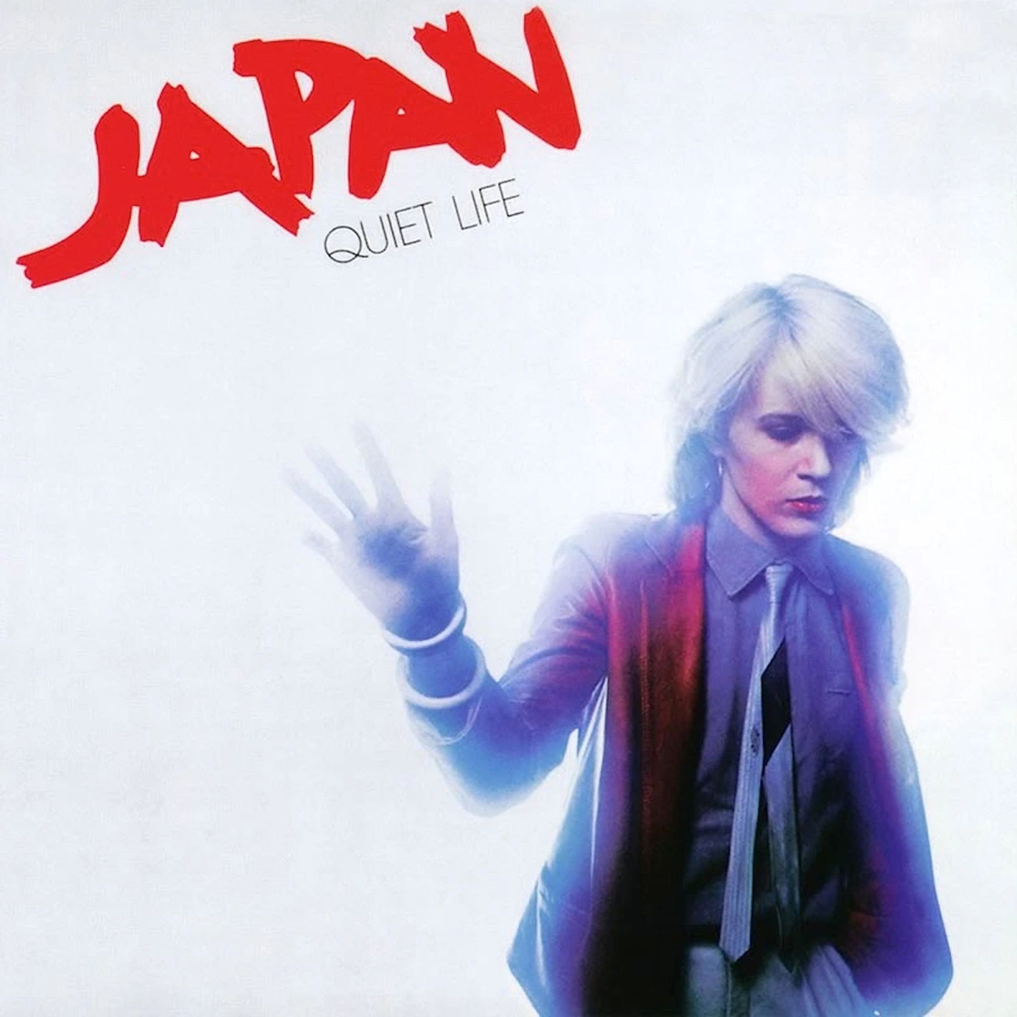 The image shows the album cover for *Quiet Life* by the band *Japan*. The cover features a minimalist design with a white background. The band’s name, "Japan," is written in bold red, stylized letters at the top left, while the album title, "Quiet Life," is placed just below in smaller, black text. The central figure is lead singer David Sylvian with light blond hair, wearing a shirt and tie, with a red jacket over it. Their hand is raised as if pushing against glass, creating a slightly blurred, ethereal effect around the edges of their figure, enhancing the moody, New Wave aesthetic.