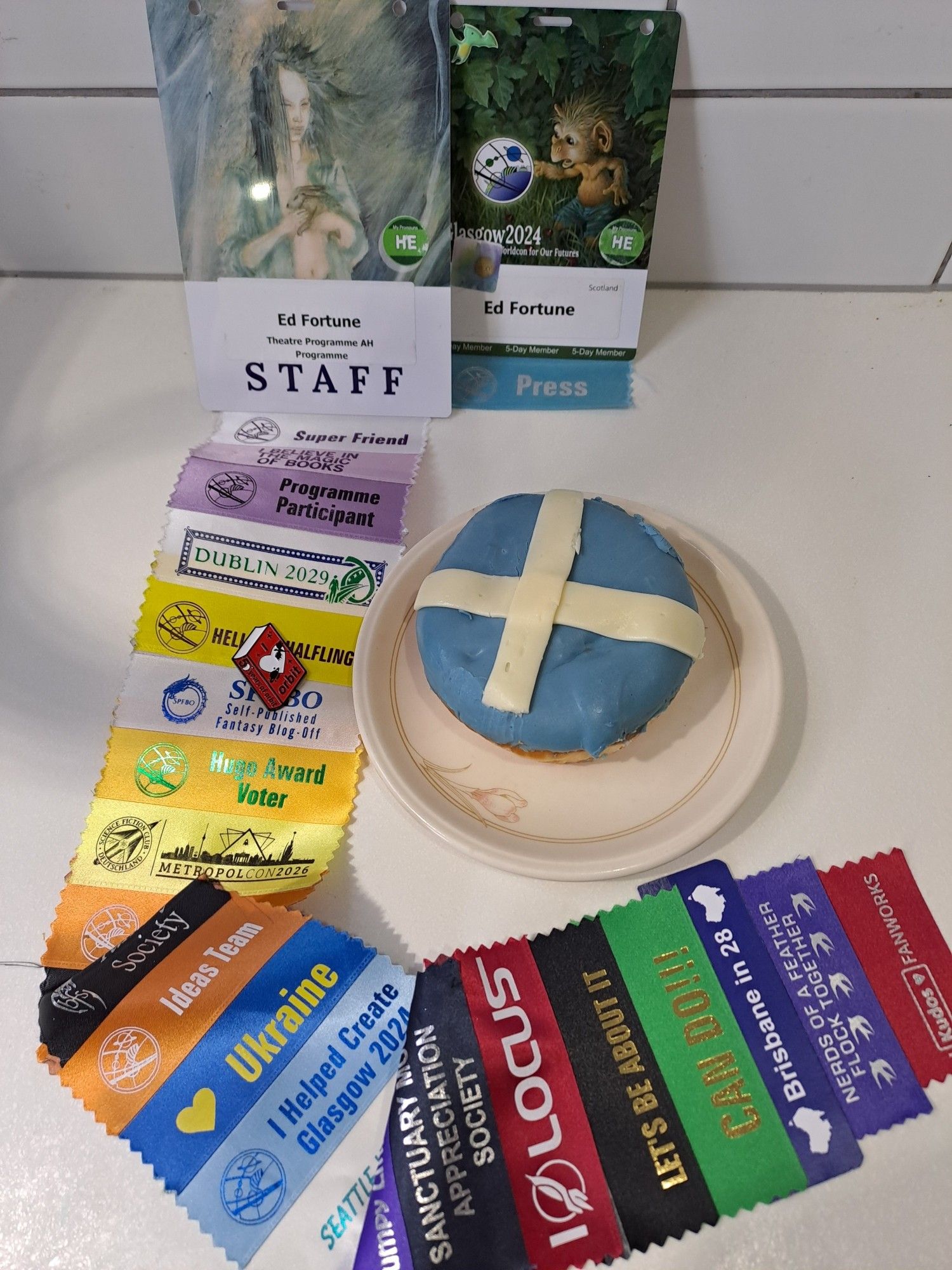 A donut with the Scottish flag design, alongside a Worldcon Staff and Press pass and many convention ribbons.