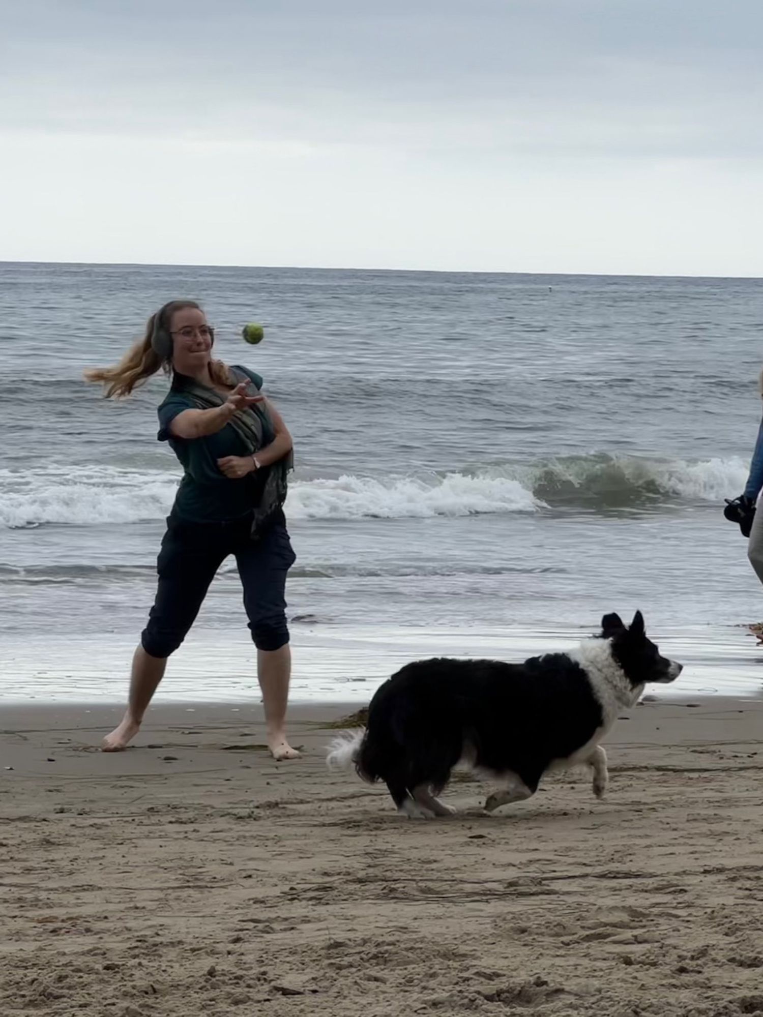 A woman (me) throwing a tennis ball for a border collie on a beach. They are just next to the water. The sky is overcast.