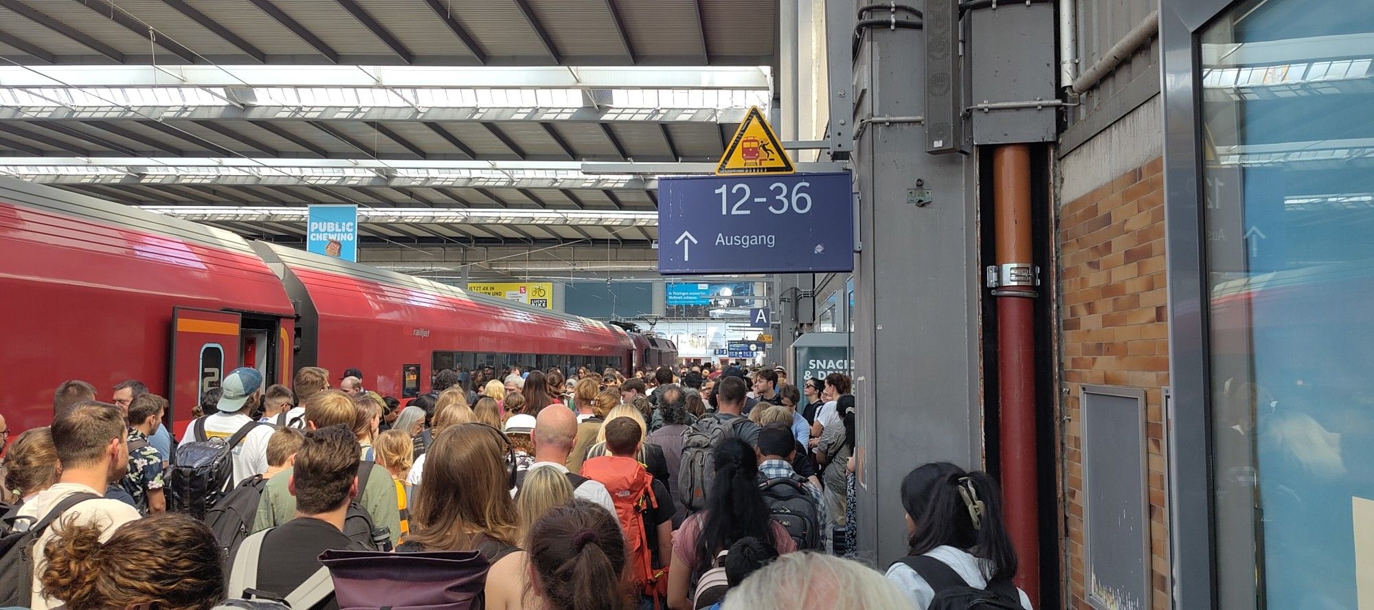 Ein mit Menschen überfüllter Bahnsteig. Links ein roter Zug, aus dem versucht wird auszusteigen. Rechts eine Wand mit Hindernissen. Der Weg ist die Verbindung zwischen den Gleisen 12-36 und 5-10