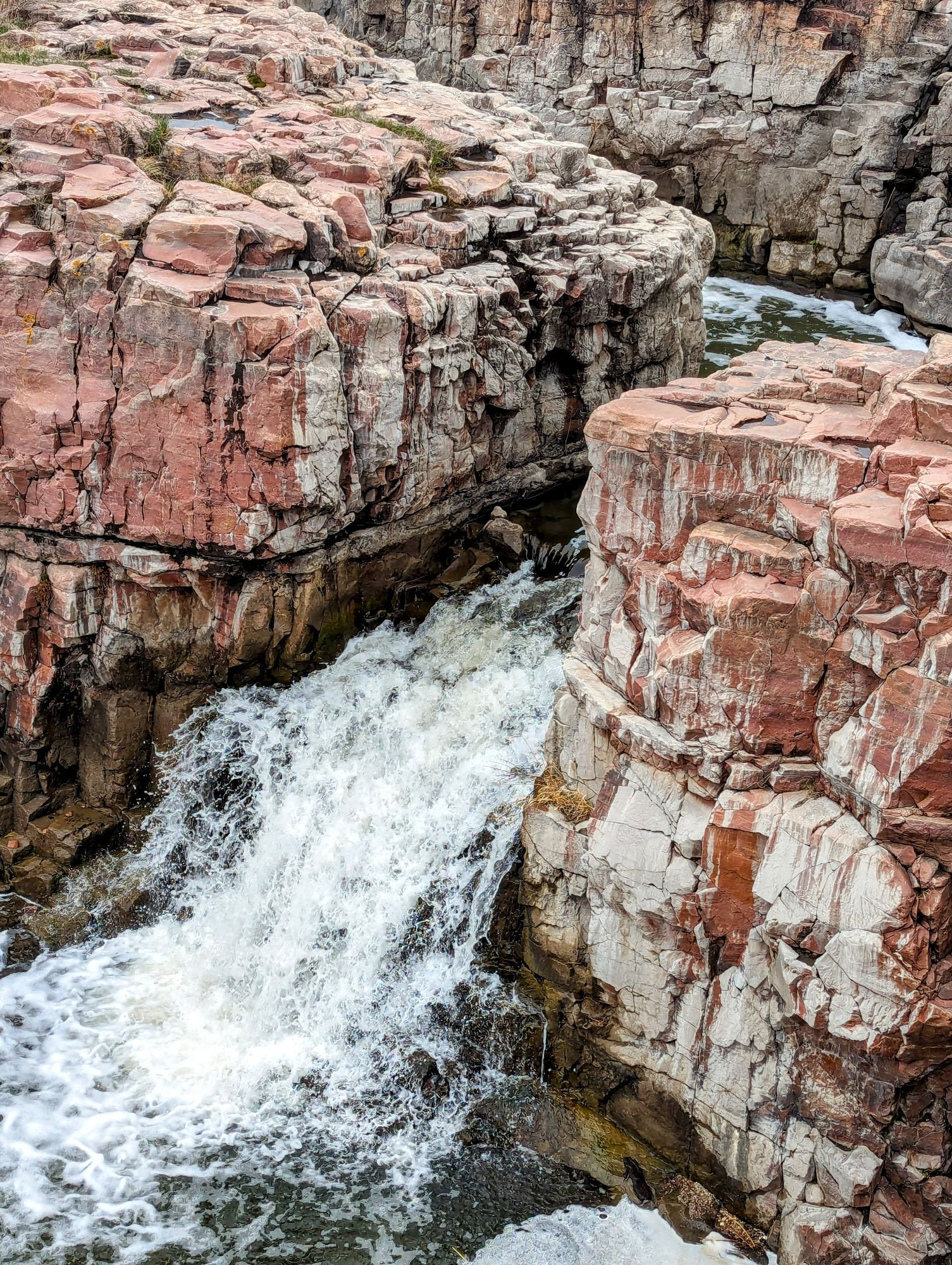 A closeup on a low set white waterfall surrounded by high red and clay colored rocks, smoothed parts jutting out in boxy shapes; surfaces from years of water flowing.