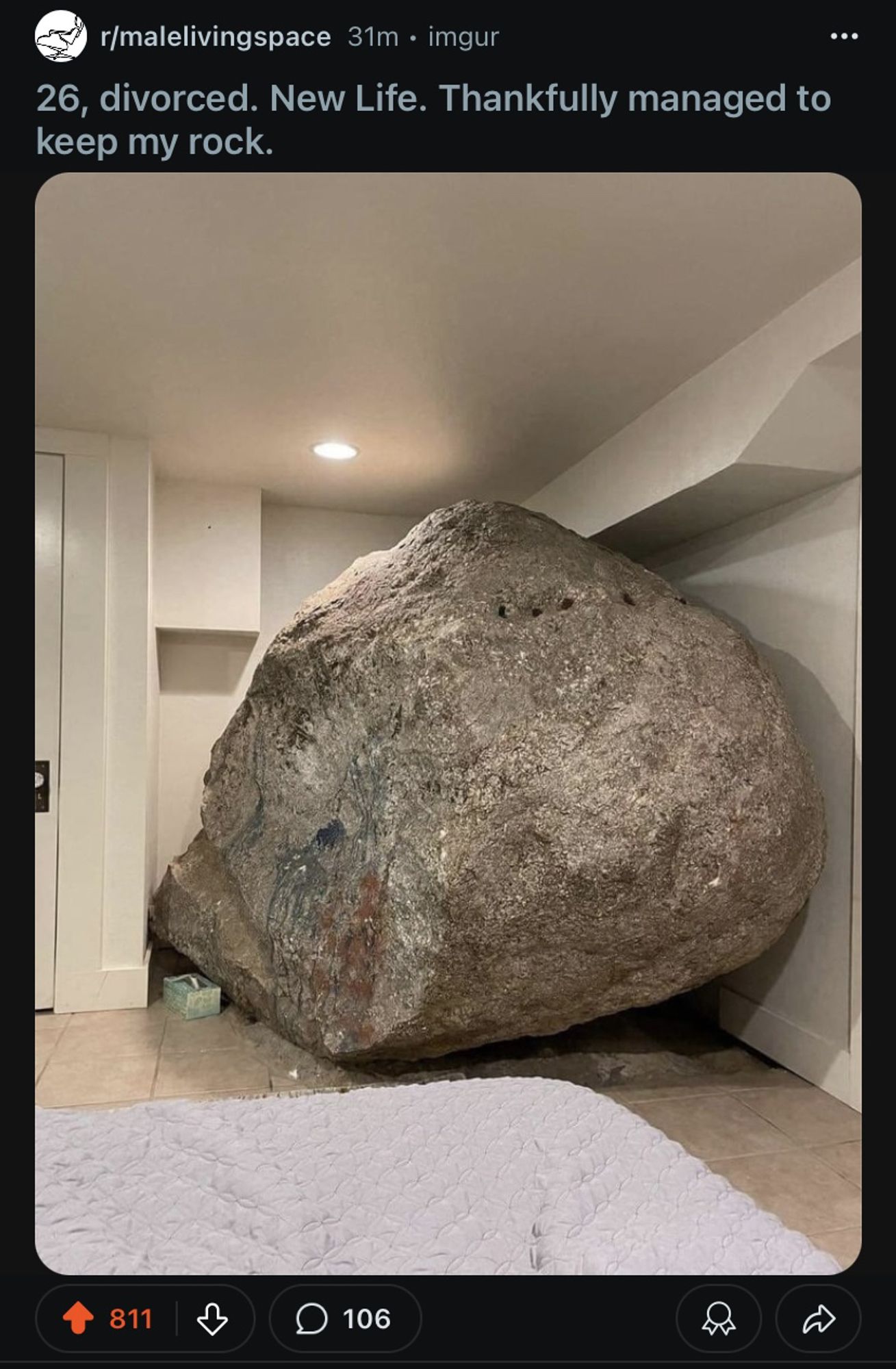 r/maleliving space post: "26, divorced. New Life. Thankfully managed to keep my rock." Above an image of what appears to be a bedroom containing an unreasonably large bolder wedged into one corner.