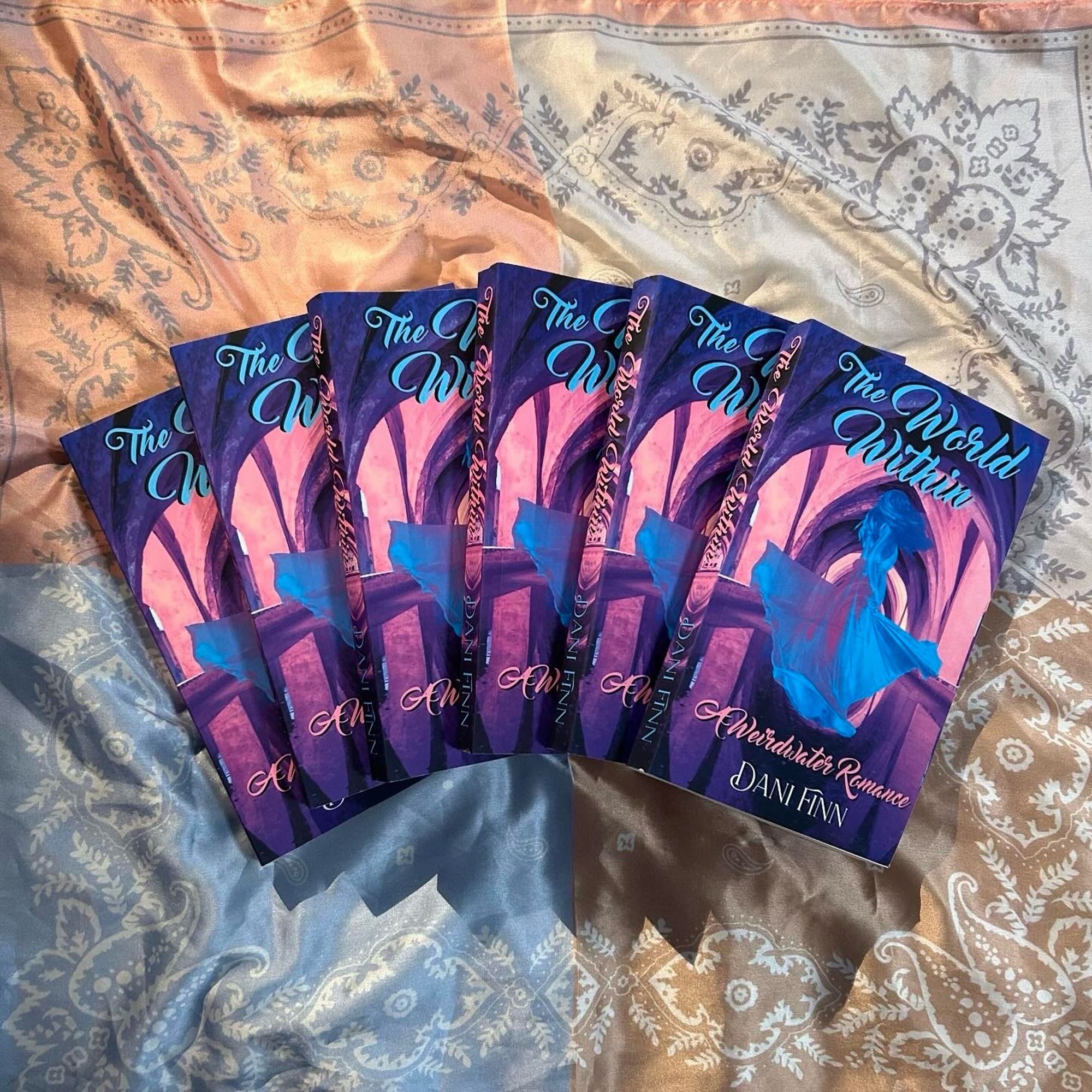 6 Copies of The World Within splayed out on a silk handkerchief. The World Within (swirly light blue font near the top), A Weirdwater Romance (swirly pink font beneath the main image) by Dani Finn (plain but slightly swirly white font near the bottom). The background image is of an arched temple pool in pink, lavender, and blue/purple tones. Floating in the center, slightly transparent, is a light blue and pinkish purple woman in a twirling dress, almost otherworldly.