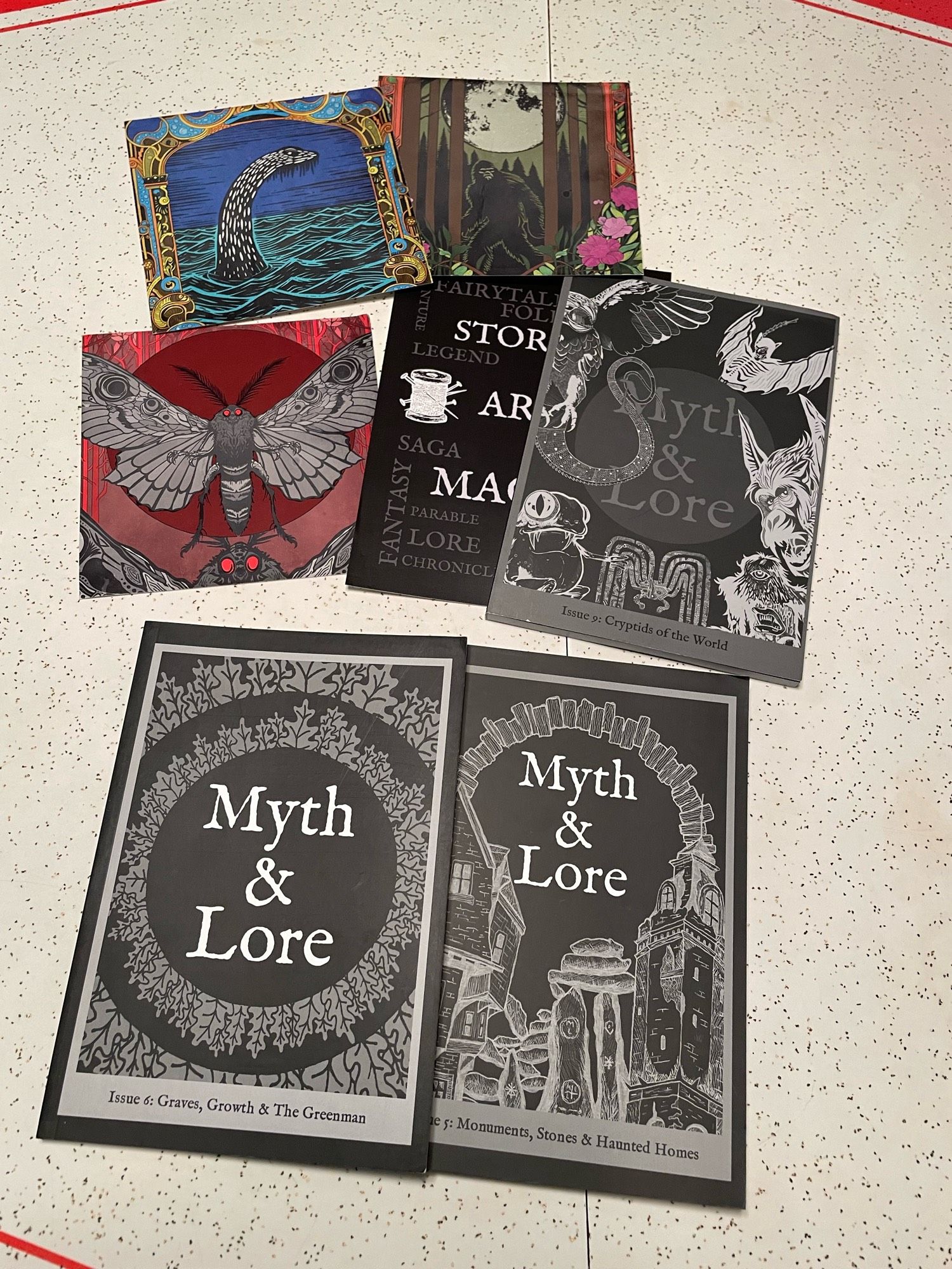 Three issues of Myth & Lore zine with black and white covers. Three full-color art prints of Mothra, Sasquatch, and Nessie