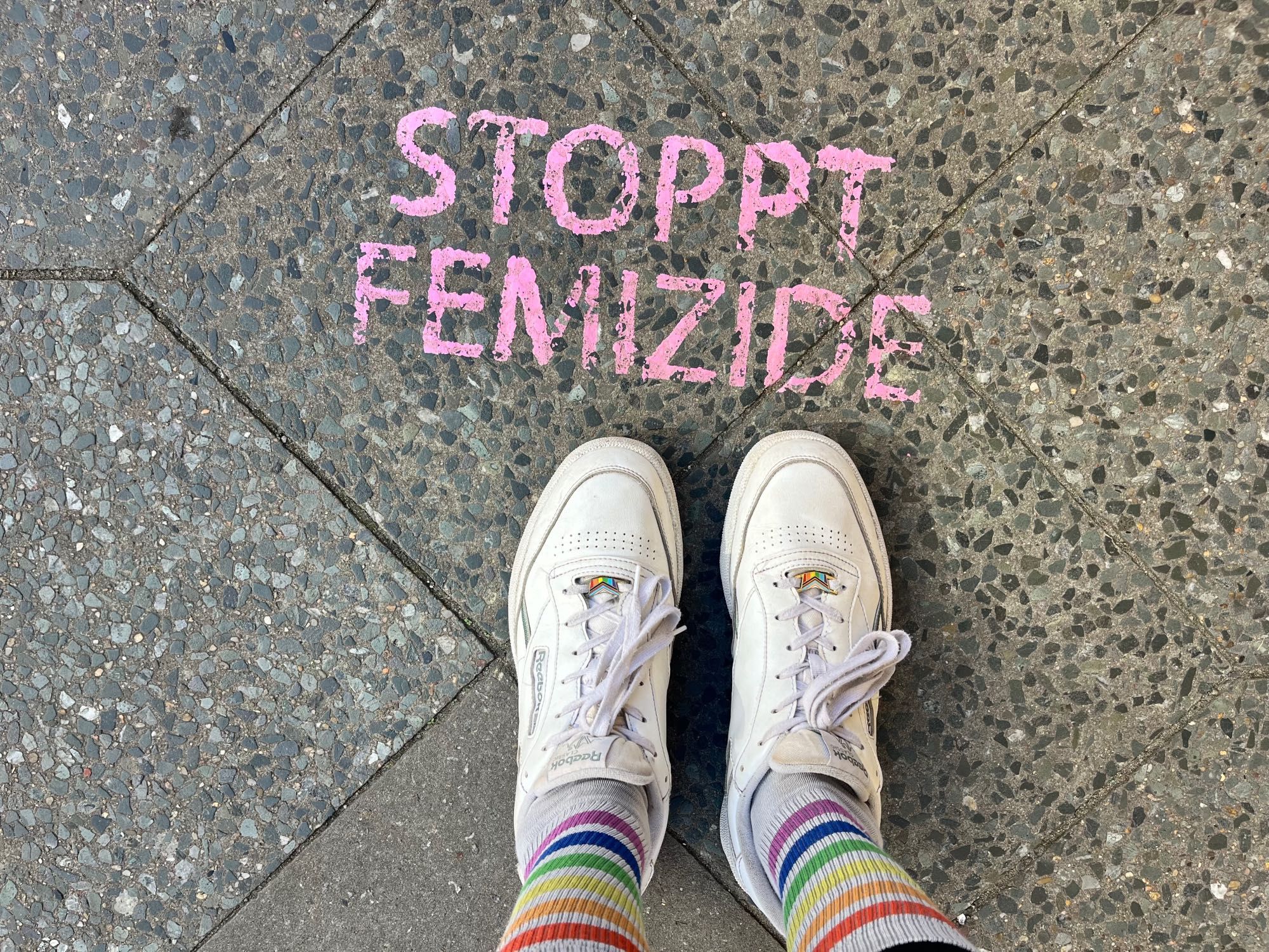 Man sieht ein paar Schuhe von oben fotografiert. Die Schuhe sind weiße Reebok-Sneaker und an den Füßen einer Person, die auch weiße Tennissocken mit Regenbogenstreifen trägt. Sie steht vor einem in rosa auf die Straße aufgesprühten „Stoppt Femizide“