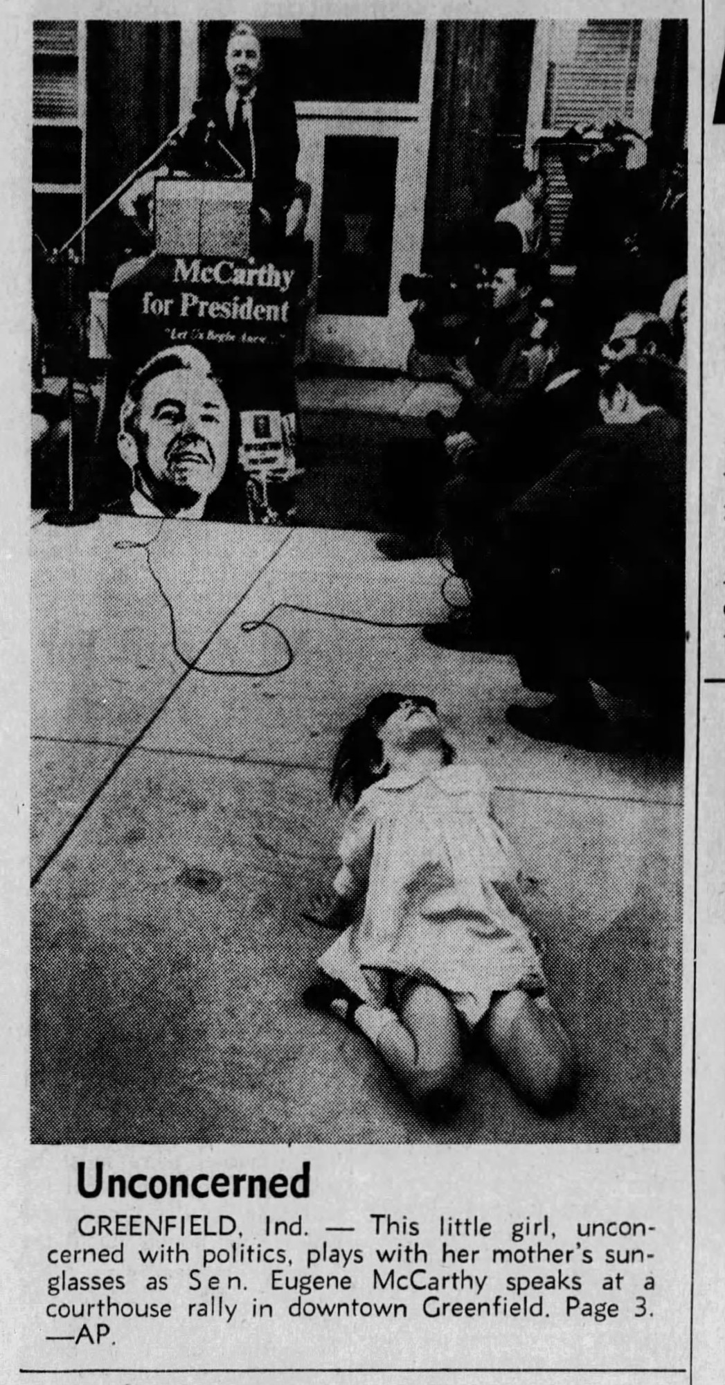 Newspaper photograph: young girl on floor in foreground and Eugene McCarthy speaking at a podium in background. Caption reads: UNCONCERNED. Greenfield, Ind.--This little girl, unconcerned with politics, plays with her mother's sunglasses as Sen. Eugene McCarthy speaks at a courthouse rally in downtown Greenfield. Page 3. --AP.