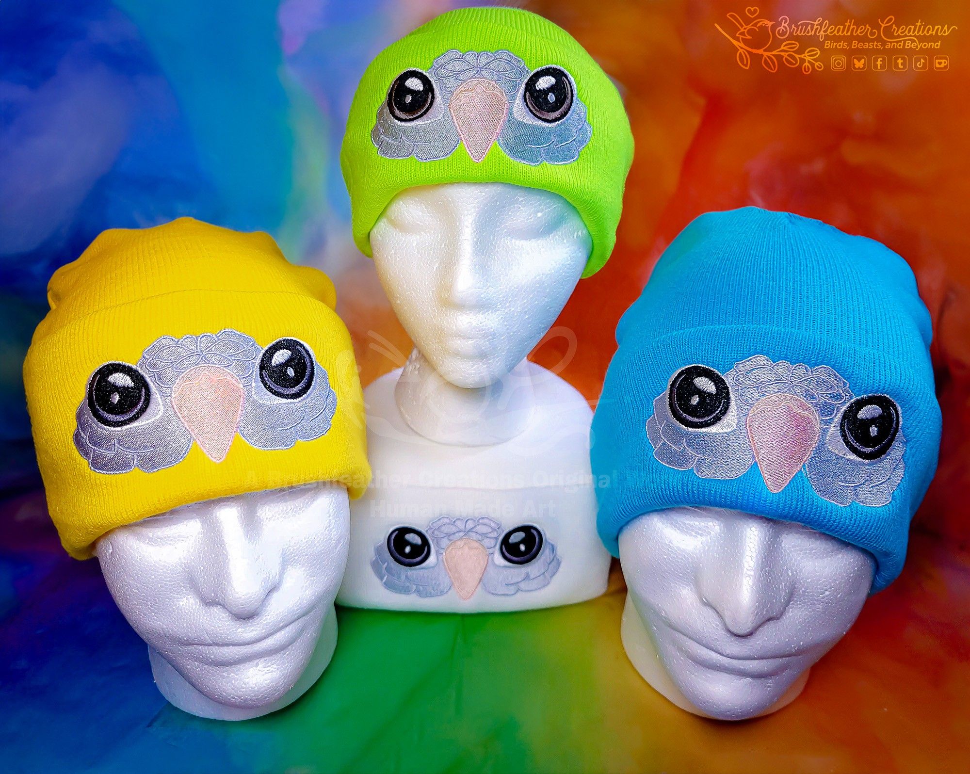 A group of 4 acrylic knit beanies on Styrofoam heads embroidered with big-eyed quaker faces in a simple, cute style. There is a yellow, a green, a white, and a blue sitting against a rainbow watercolor background.