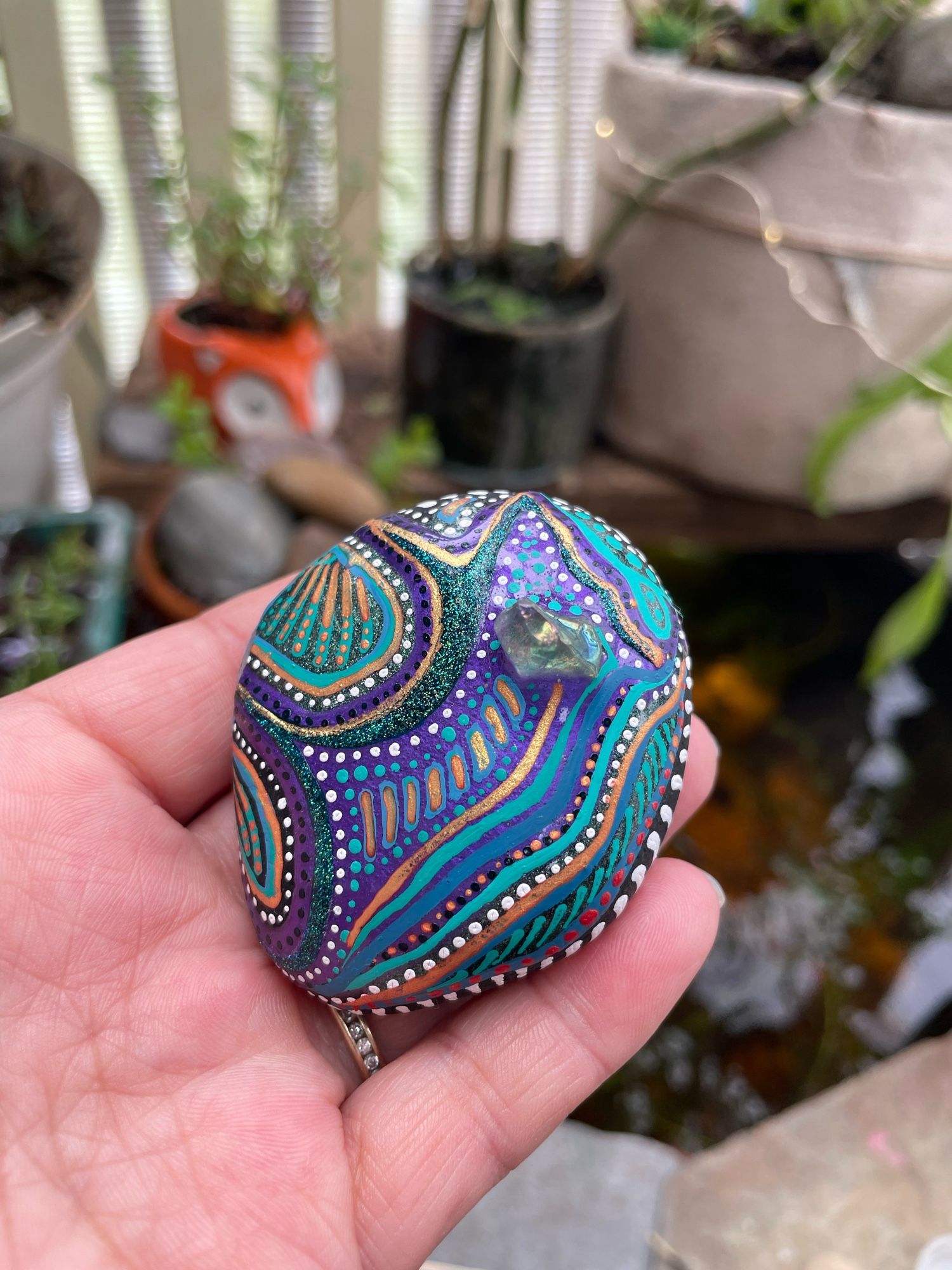 A hand holds a rock that has been painted in blues, purples, and greens. The lines and dots compliment the shape of the rock. A single piece of tumbled glass stone is attached as well