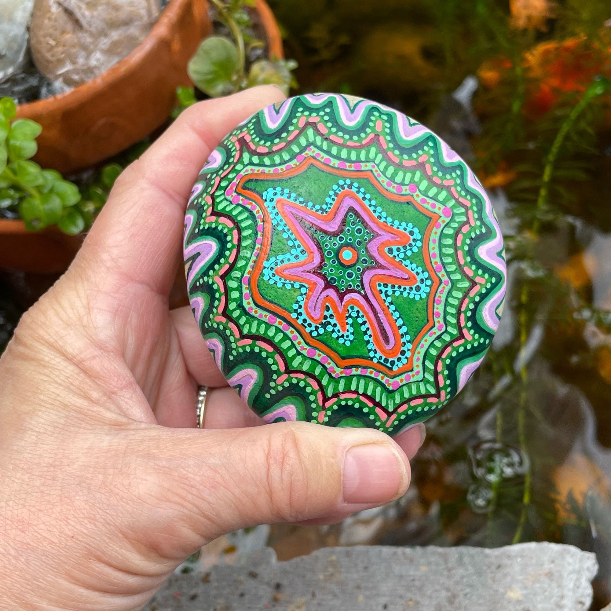 My hand holding a a rock painted in wavy lines of pinks, blues and greens, accented by dots and lines