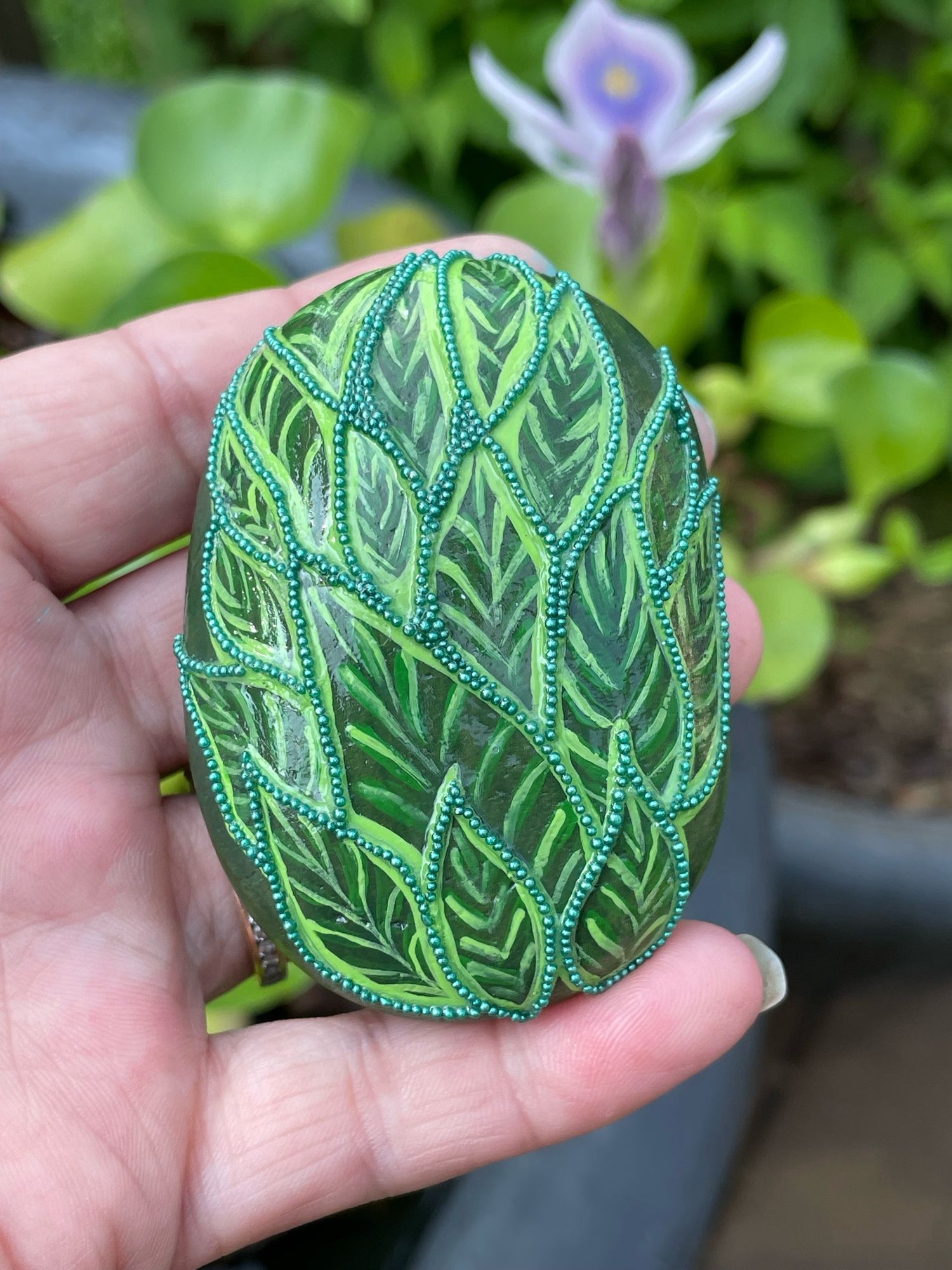 I’m holding a painted rock that is green. It has been made to look like leaves and the edges are highlighted with green pixie beads.