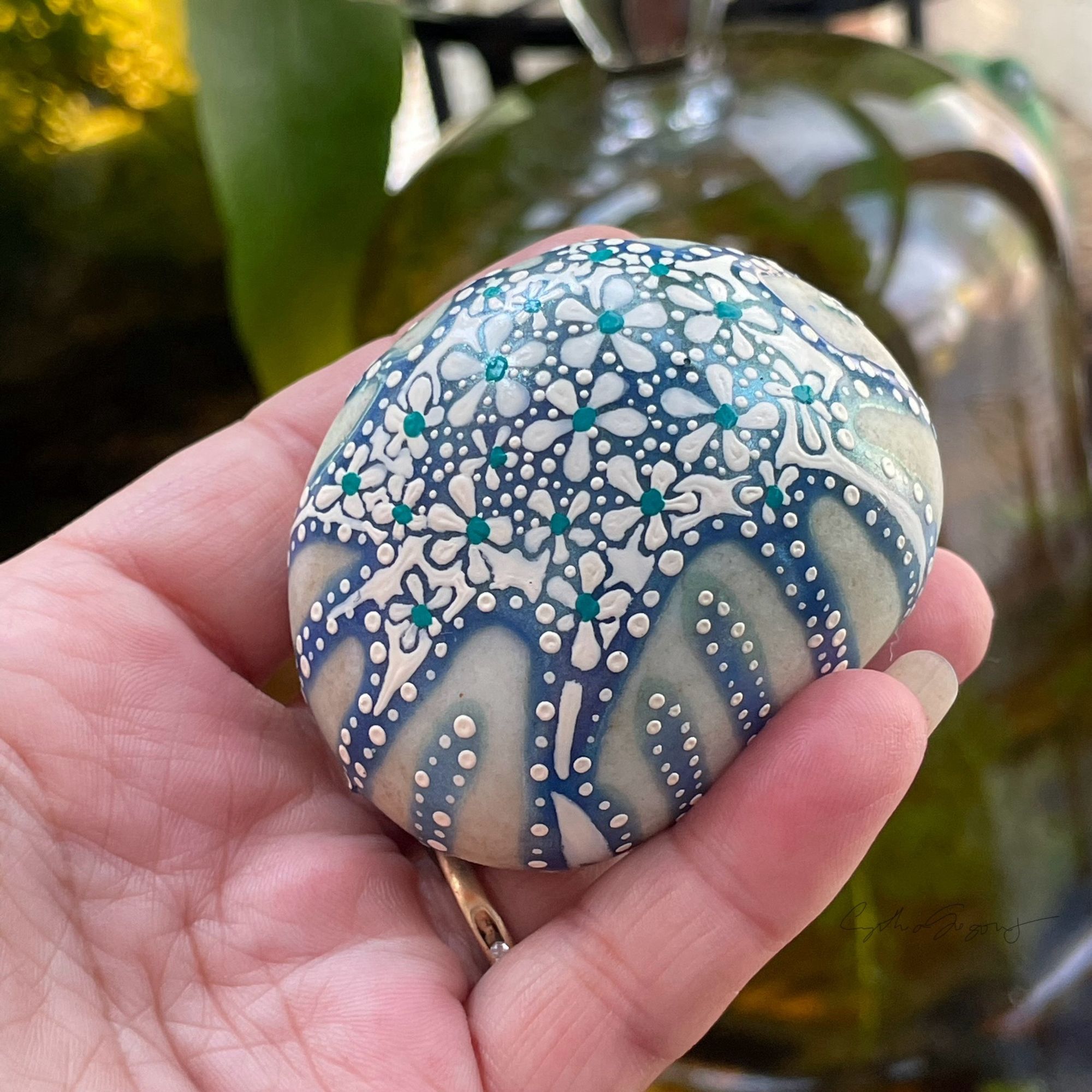 This photo is focused on a white rock with a dripping blue and white flower design. The design is a simple 5-petal, white flower with a teal dot center. This is repeated in different sizes in random, but comfortable placent.