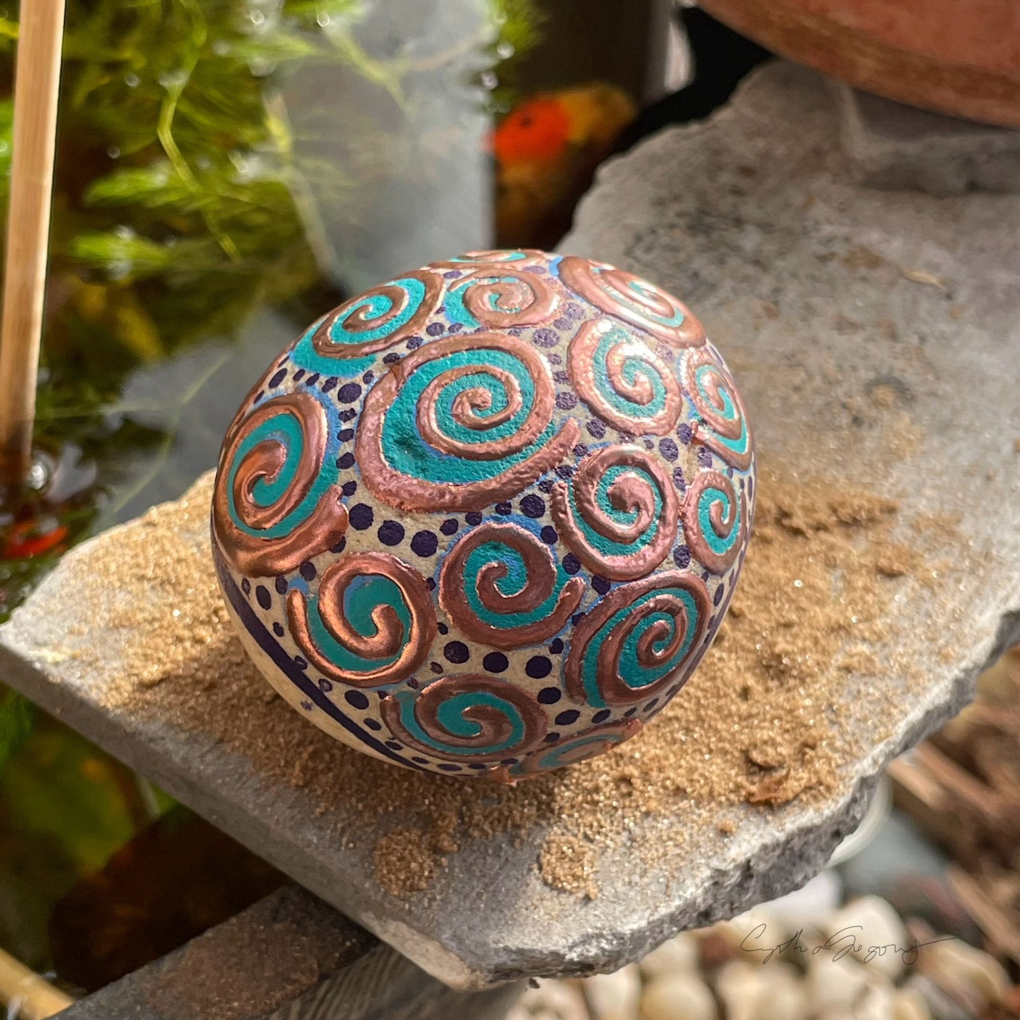 Angled view down at a tan rock adorned with teal spiral shapes highlighted with raised copper spirals. Dark blue dots fill many blank spaces.