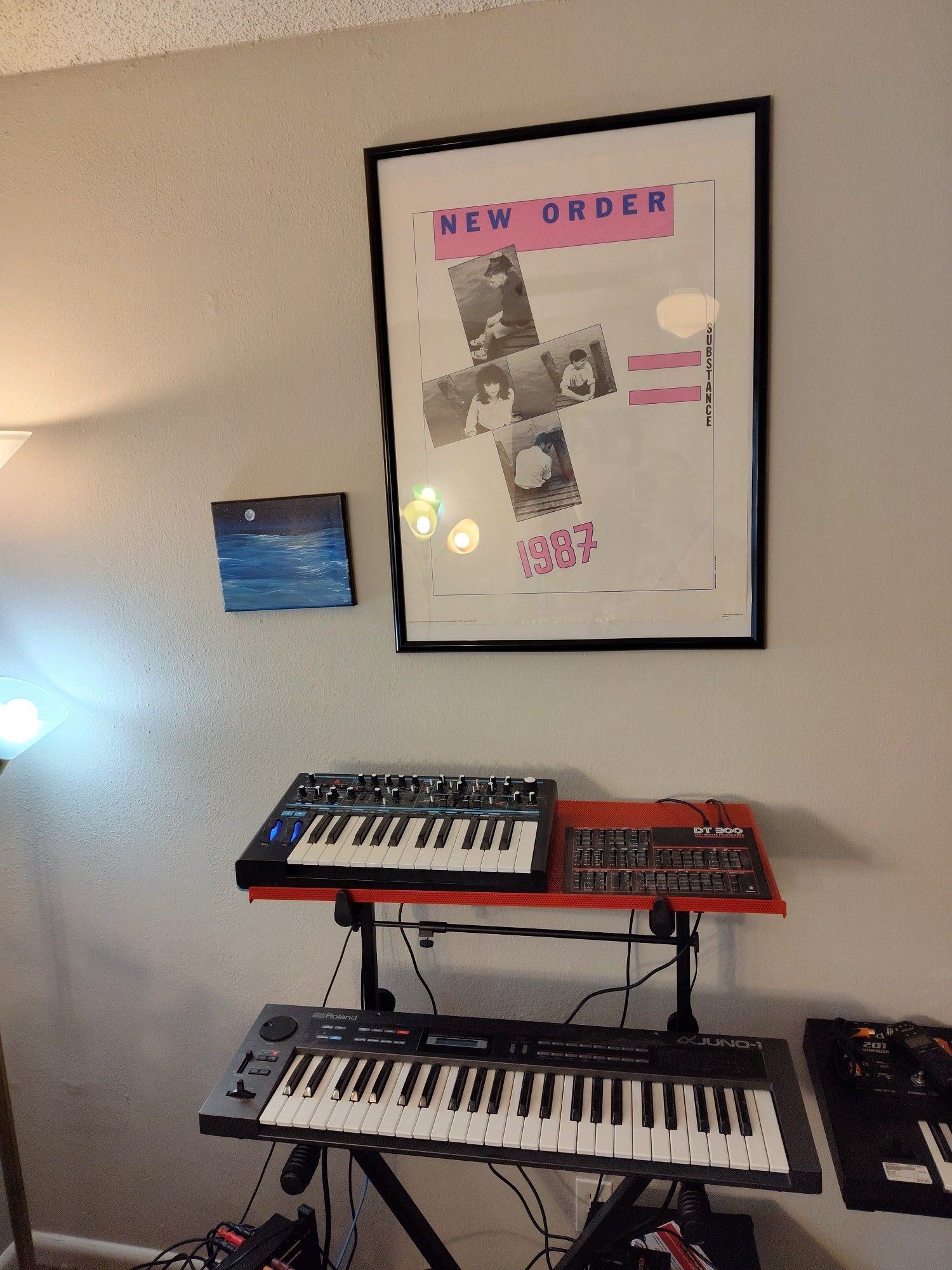 A photo of my music studio, featuring a vintage poster for the band New Order and a painting of a body of water at night situated on my wall above a set of synths