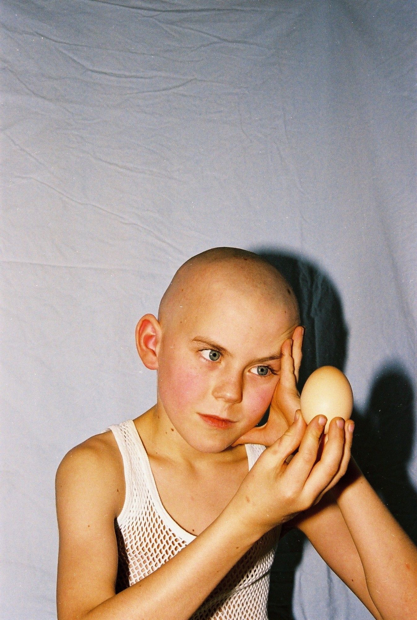 A seven year old bald boy in a tanktop, staring at an egg.