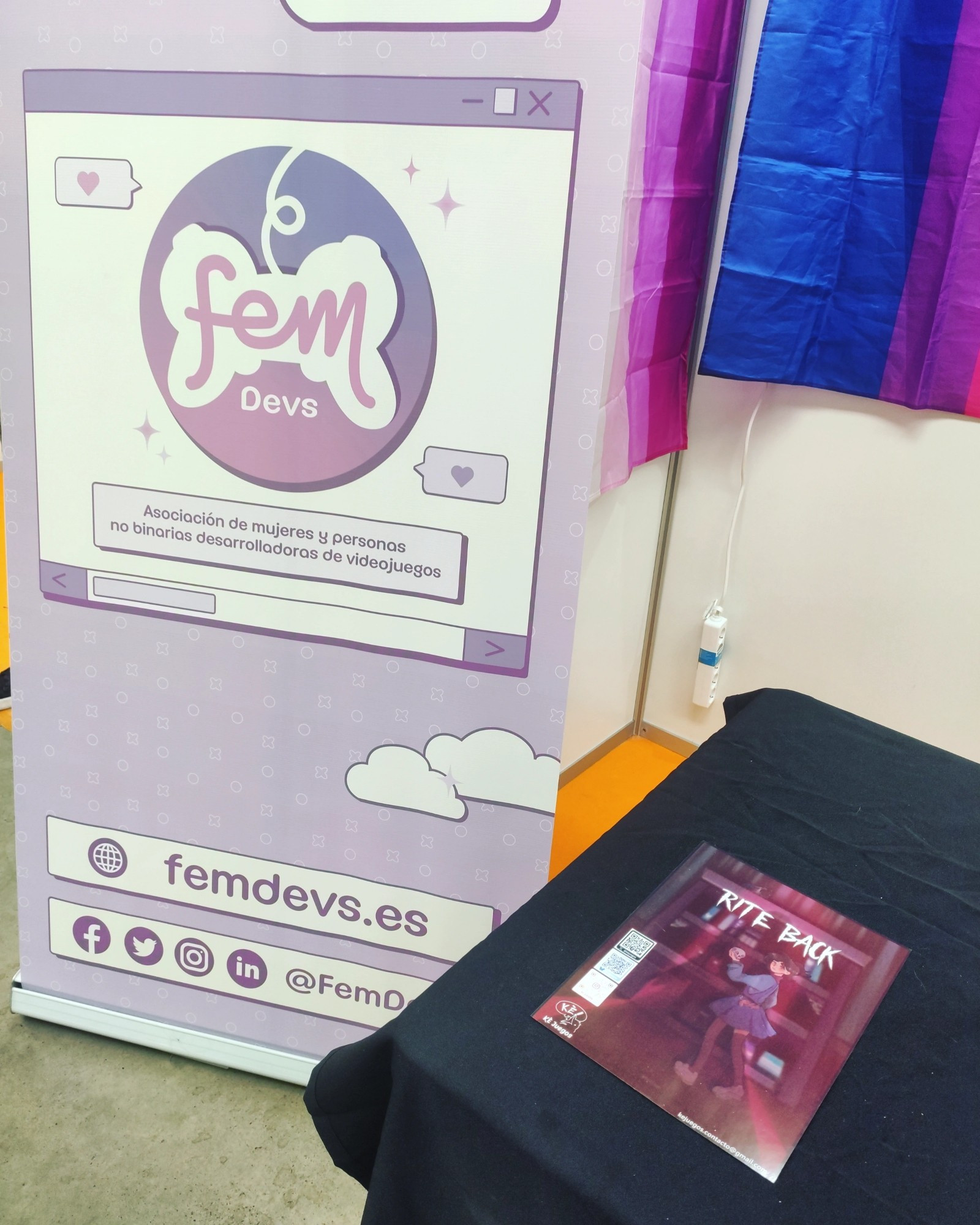 KÉ Juegos en el stand de Femdevs en la IndieDevDay, poster de Rite Back encima de la mesa y roll up de femdevs