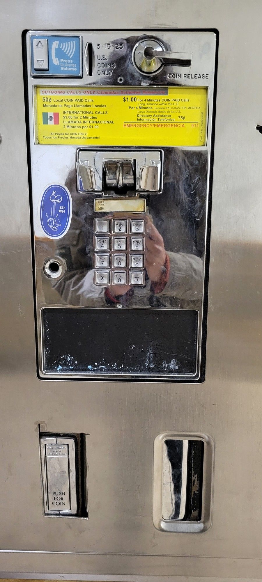 A payphone still with information on cost per minute in English and Spanish. The receiver removed. You can see the reflection of my hand on the surface, phone hidden behind the number buttons.